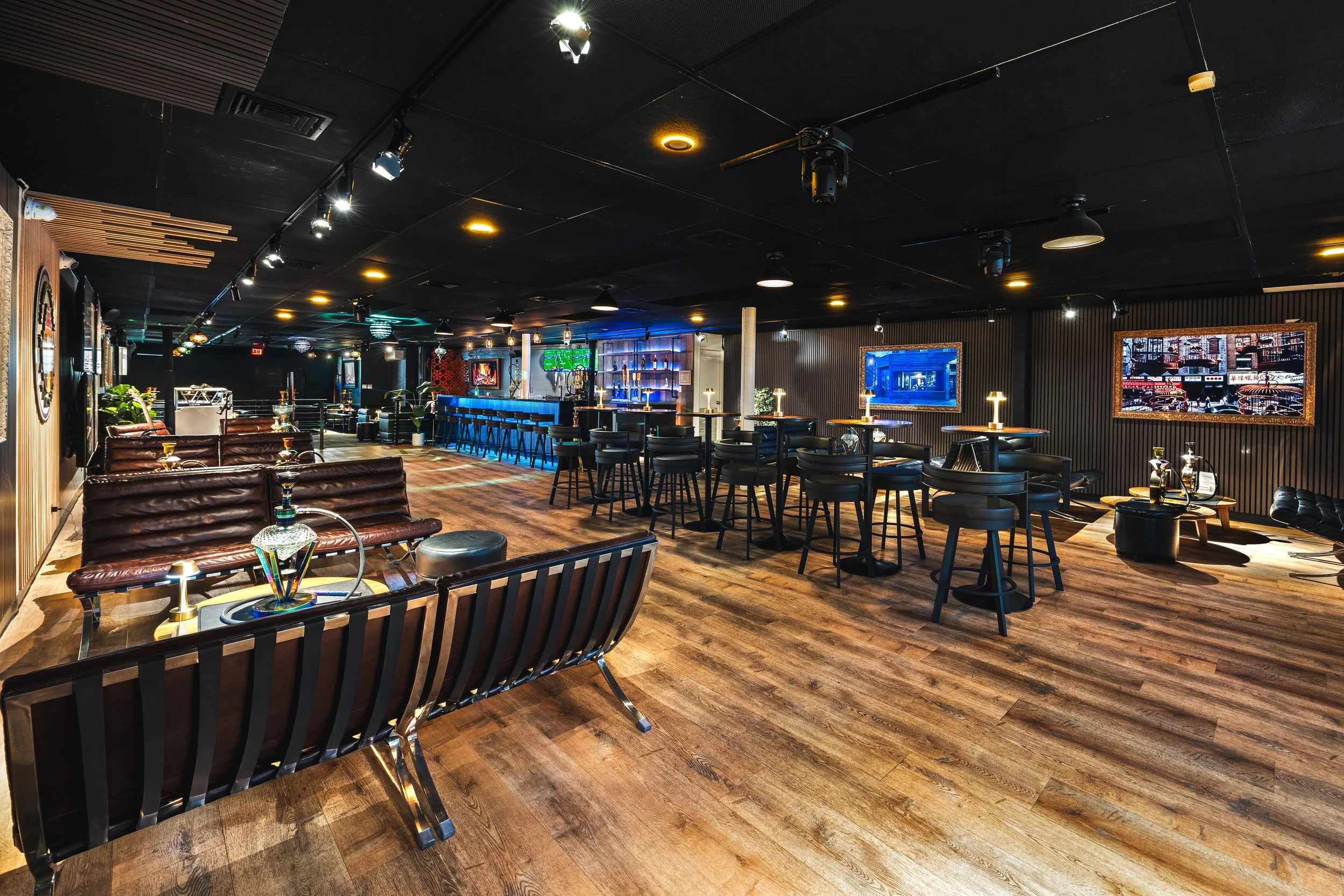 Interior of a modern bar with wooden flooring, black ceiling with spotlights, and dark walls. There are leather couches and tables with hookahs, a bar counter with high chairs, and illuminated screens on the walls.