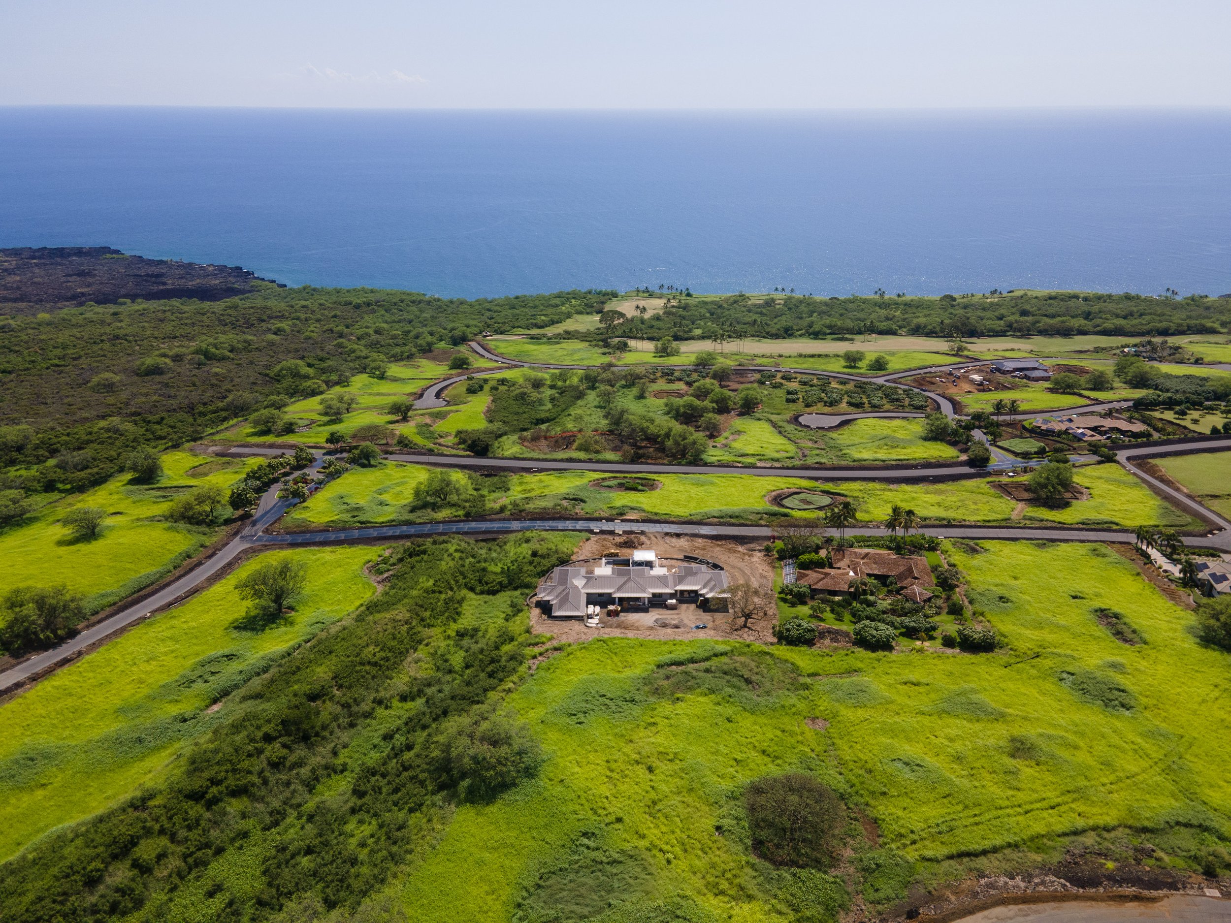 Aerial view of lush green farmland with winding roads, houses, and trees overlooking the ocean in the distance.