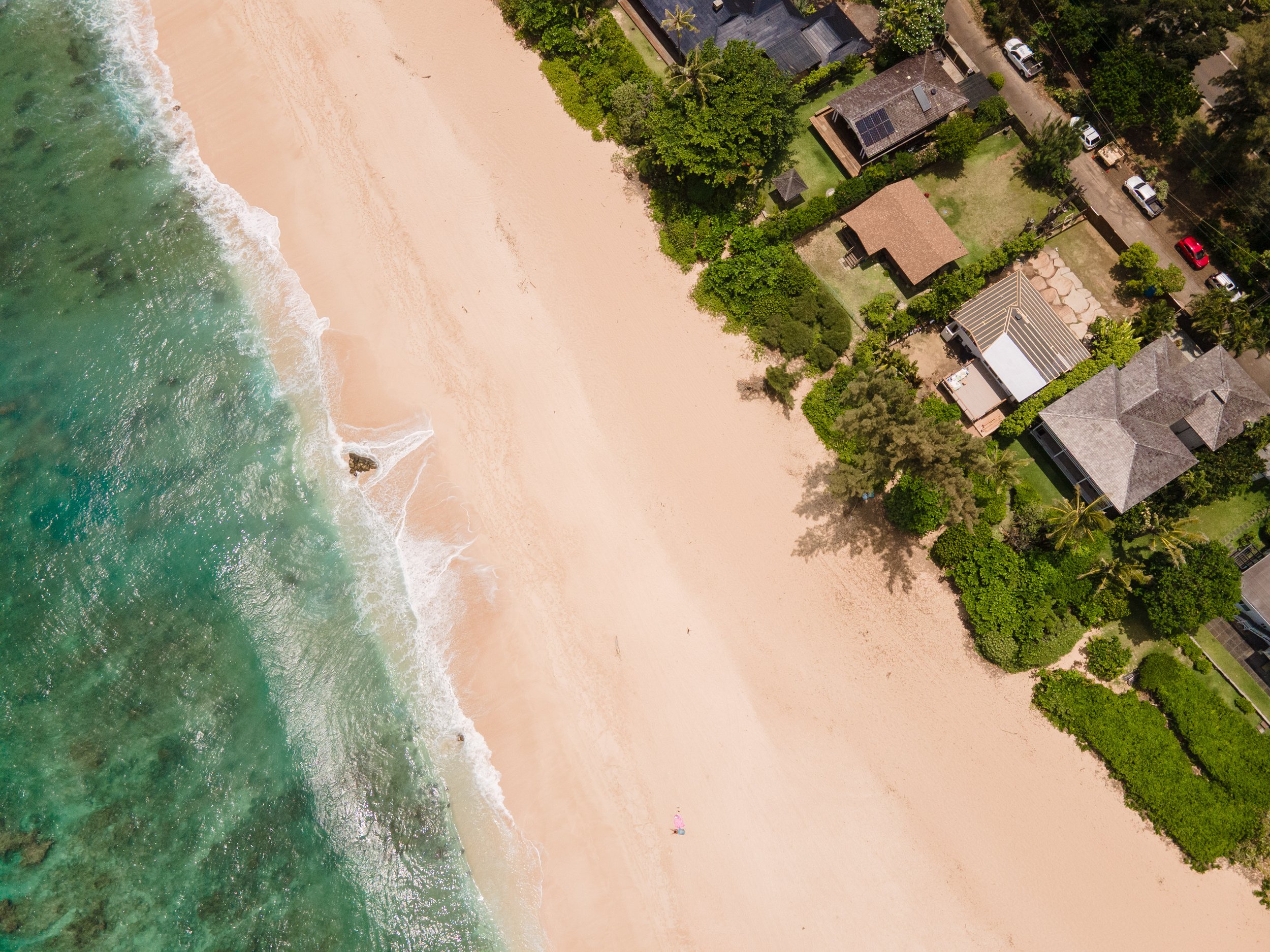 An aerial view of a beach with clear greenish water, white sand, and greenery next to residential houses with driveways, parked cars, and trees.