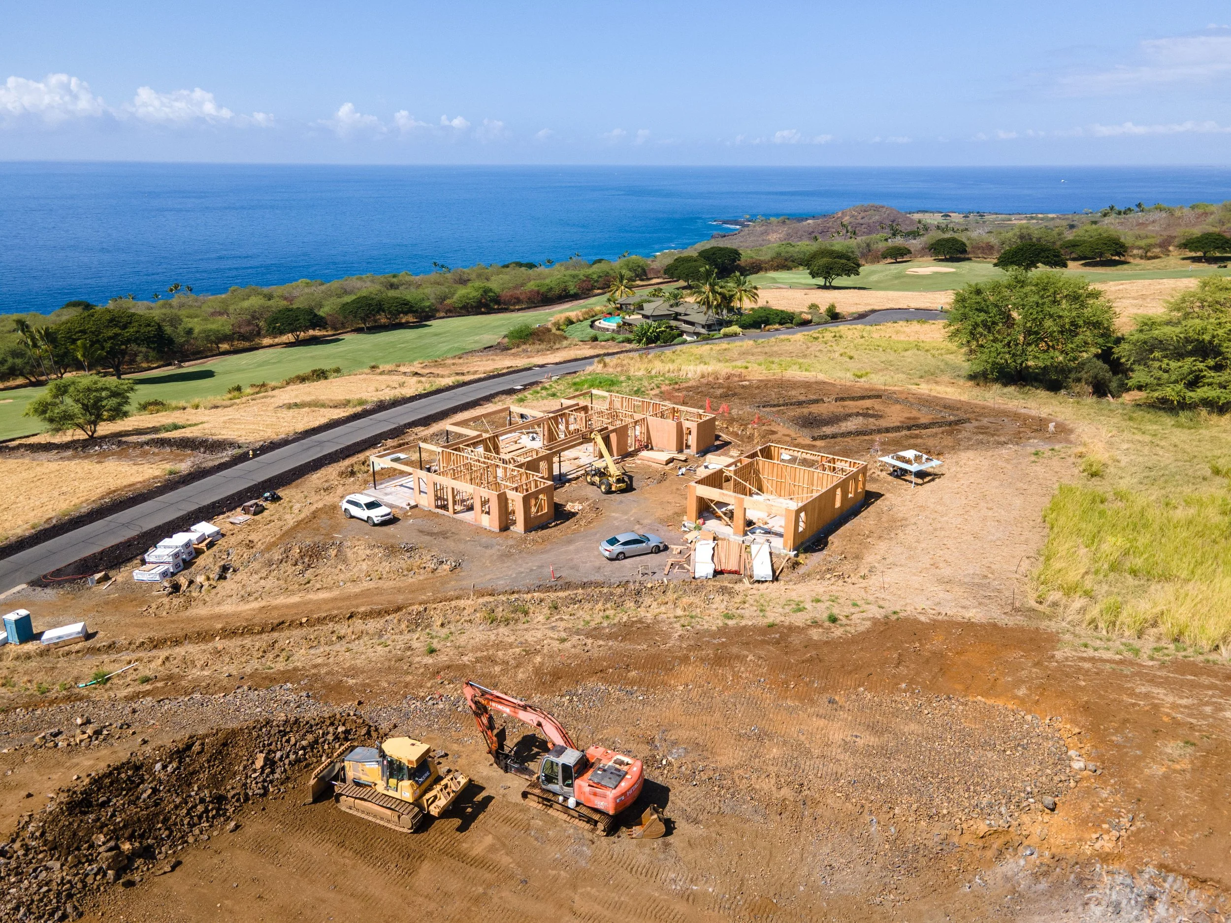 Construction site with building framing, construction vehicles, and machinery near a coastal landscape with trees, a golf course, and the ocean in the background.