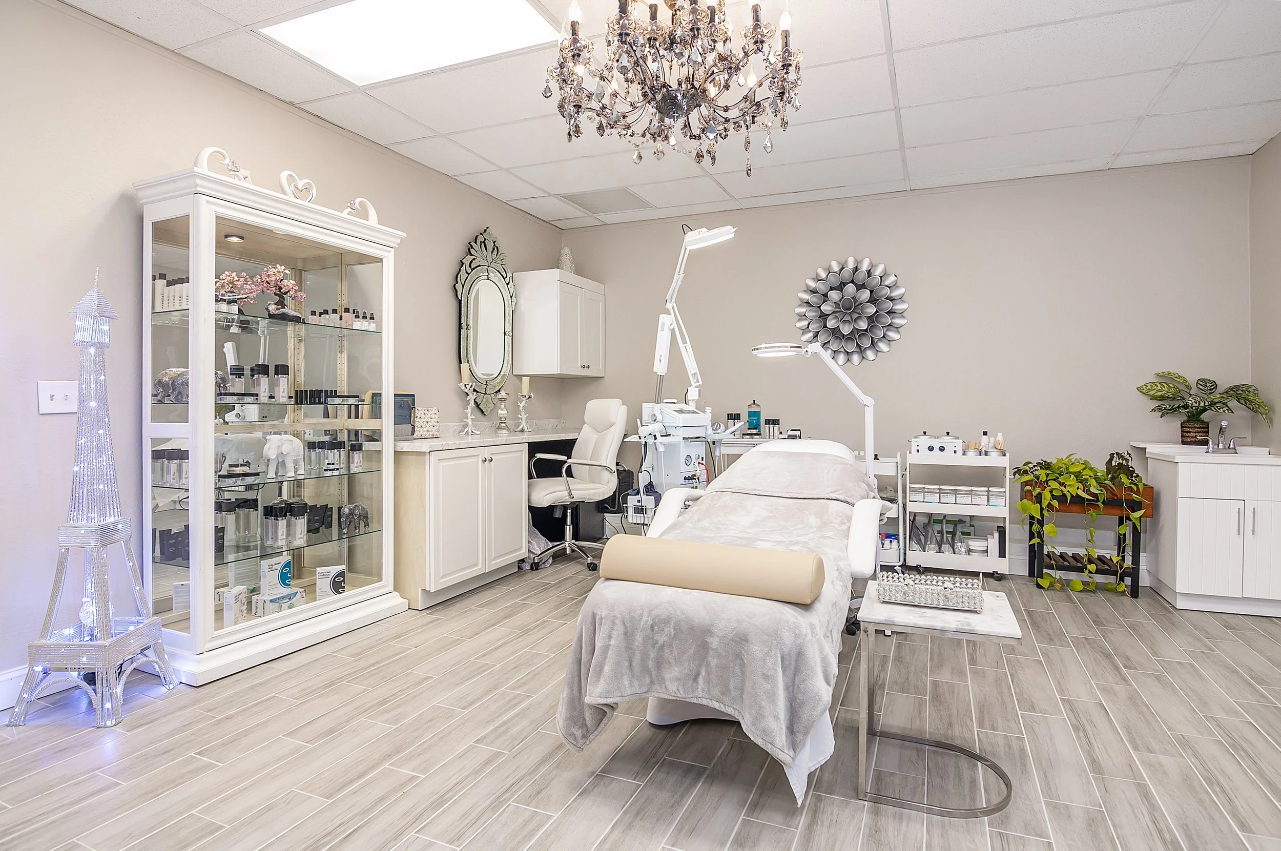 A medical or beauty treatment room with a treatment bed, robotic equipment, and storage cabinets. Decor includes a chandelier, a decorative wall piece, and plants.