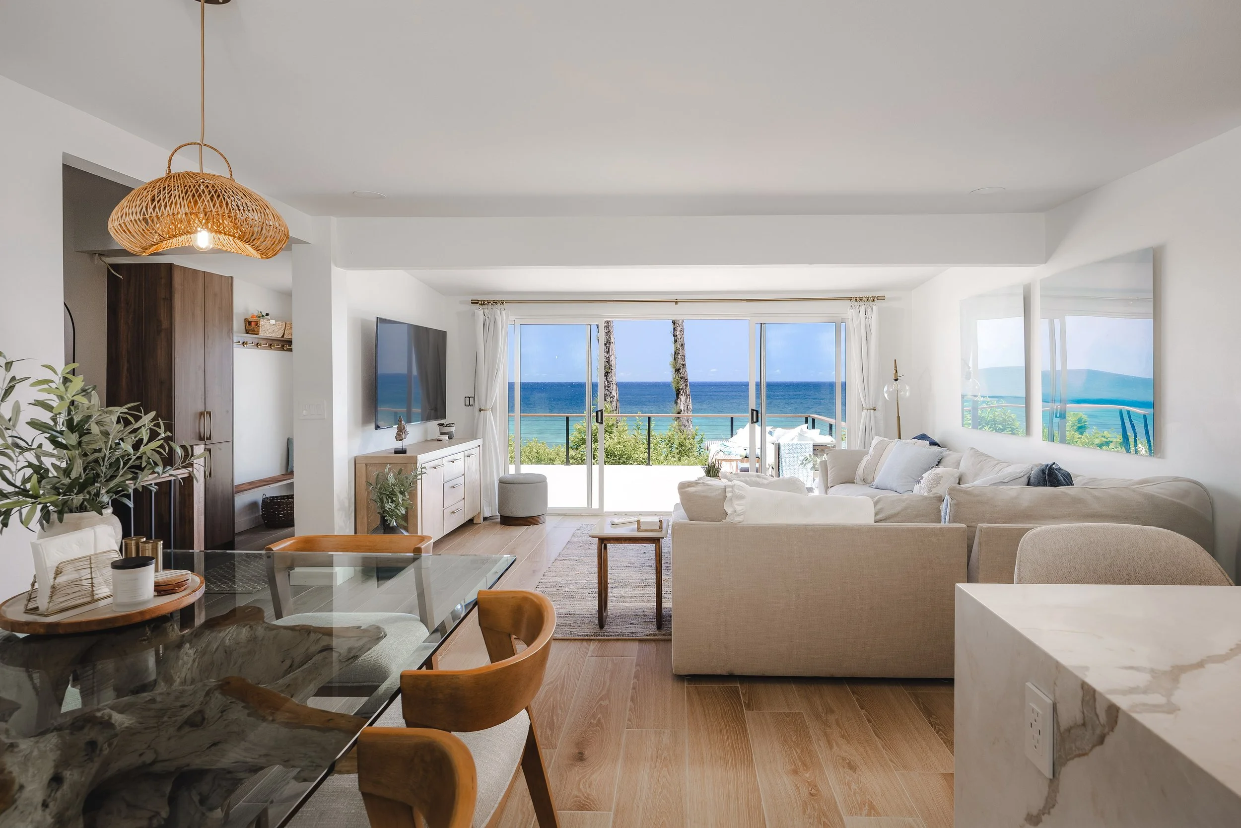 Living room with large glass sliding doors showing the ocean view, white walls, beige couches, wooden coffee table, flat-screen TV, and wicker pendant light.