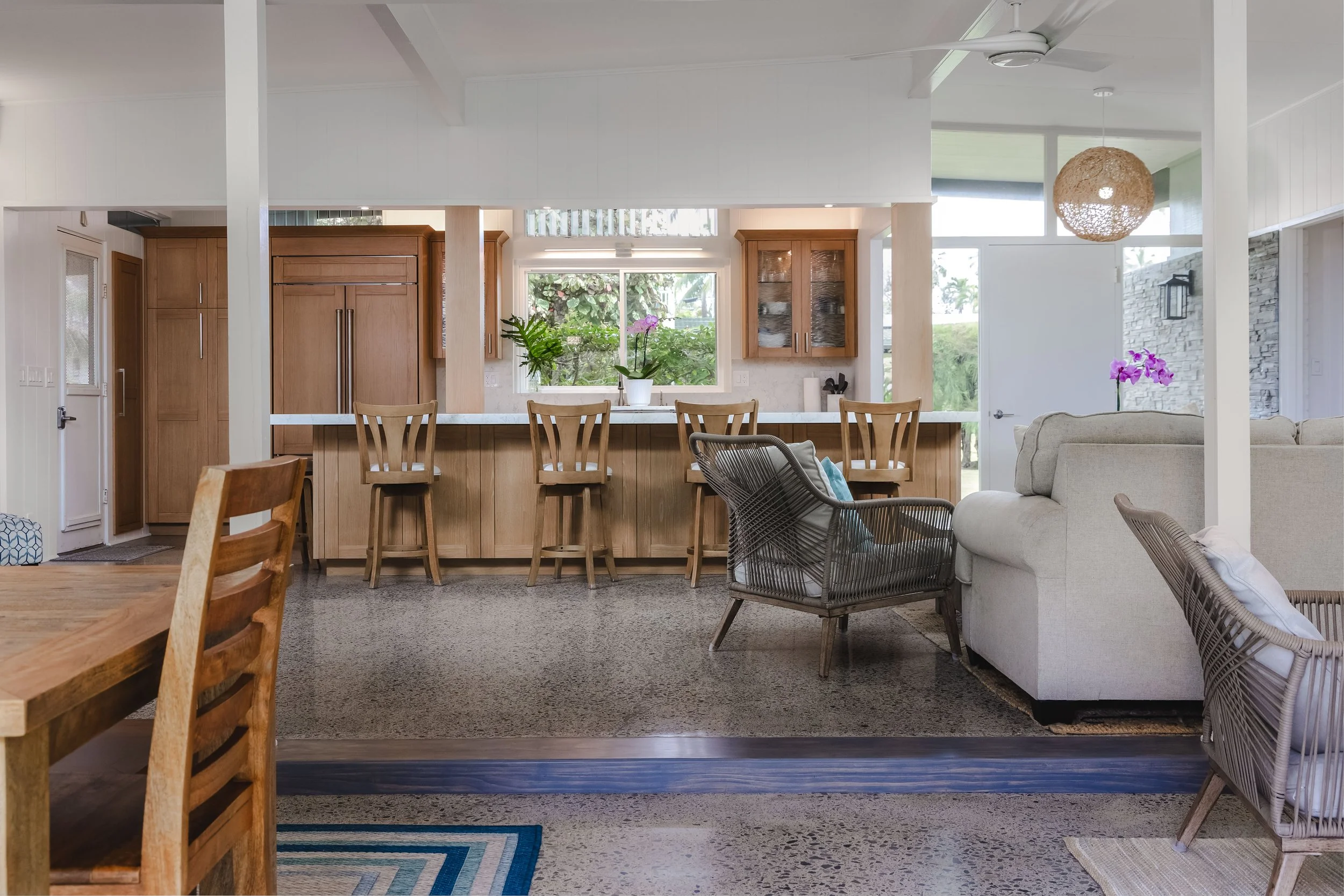 A view of a modern open kitchen and living room with wooden cabinets, a white countertop, potted plants, and natural light from large windows and glass doors.
