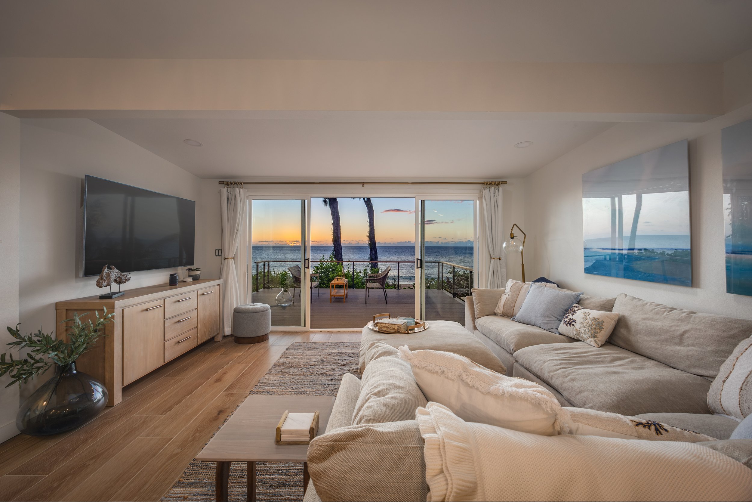 Living room with large sliding glass doors opening to a balcony with ocean view at sunset. Interior includes a beige sectional sofa, a wooden sideboard with decorative items, a flat-screen TV, a glass chandelier, and a patterned rug.