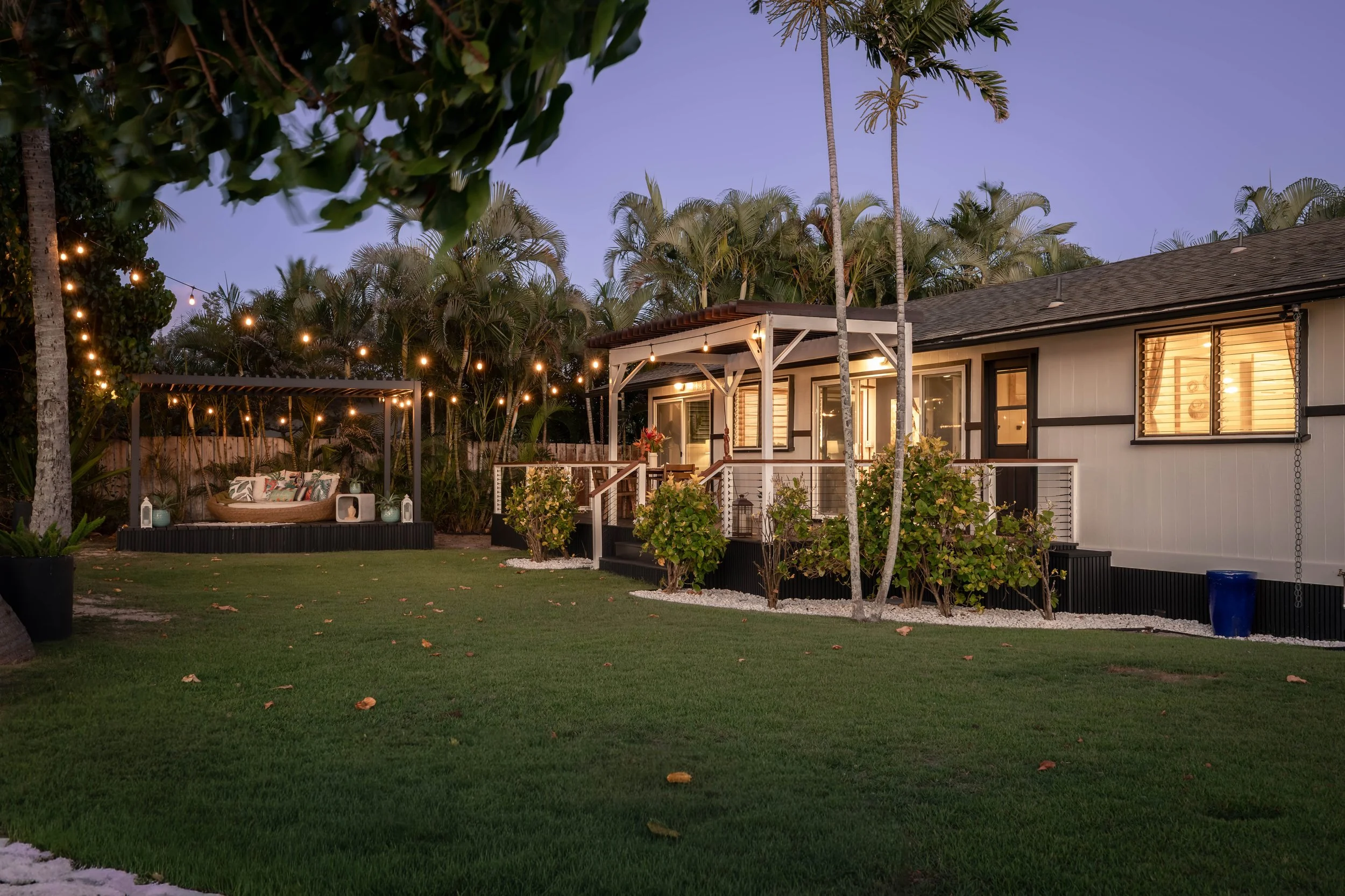 A backyard scene during dusk with a small house, palm trees, string lights, a patio area, and well-maintained lawn.