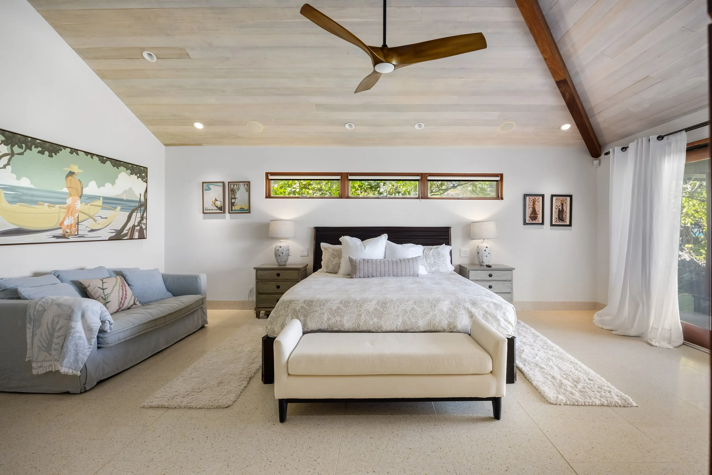 A bedroom with a large bed in the center, flanked by two nightstands with lamps. There is a gray sofa with pillows on the left, a painting on the wall beside it, and decorative art pieces on the right wall. The room has a wooden ceiling fan, horizont
