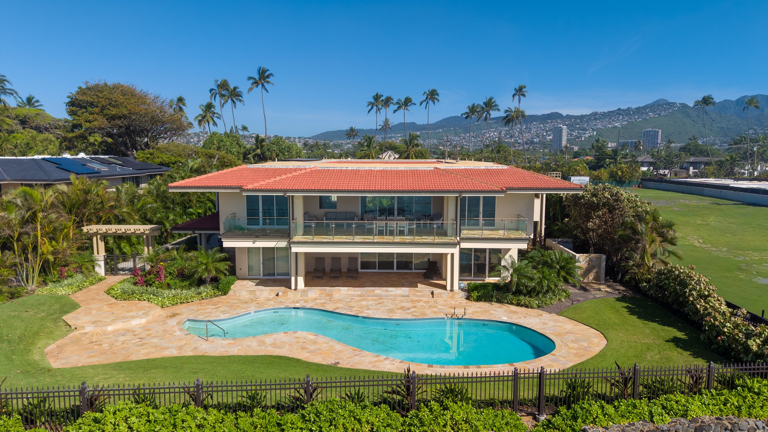 A two-story house with a red-tiled roof, large glass windows, and a balcony, surrounded by a lush green garden with a swimming pool in the backyard, with palm trees and mountains in the background.