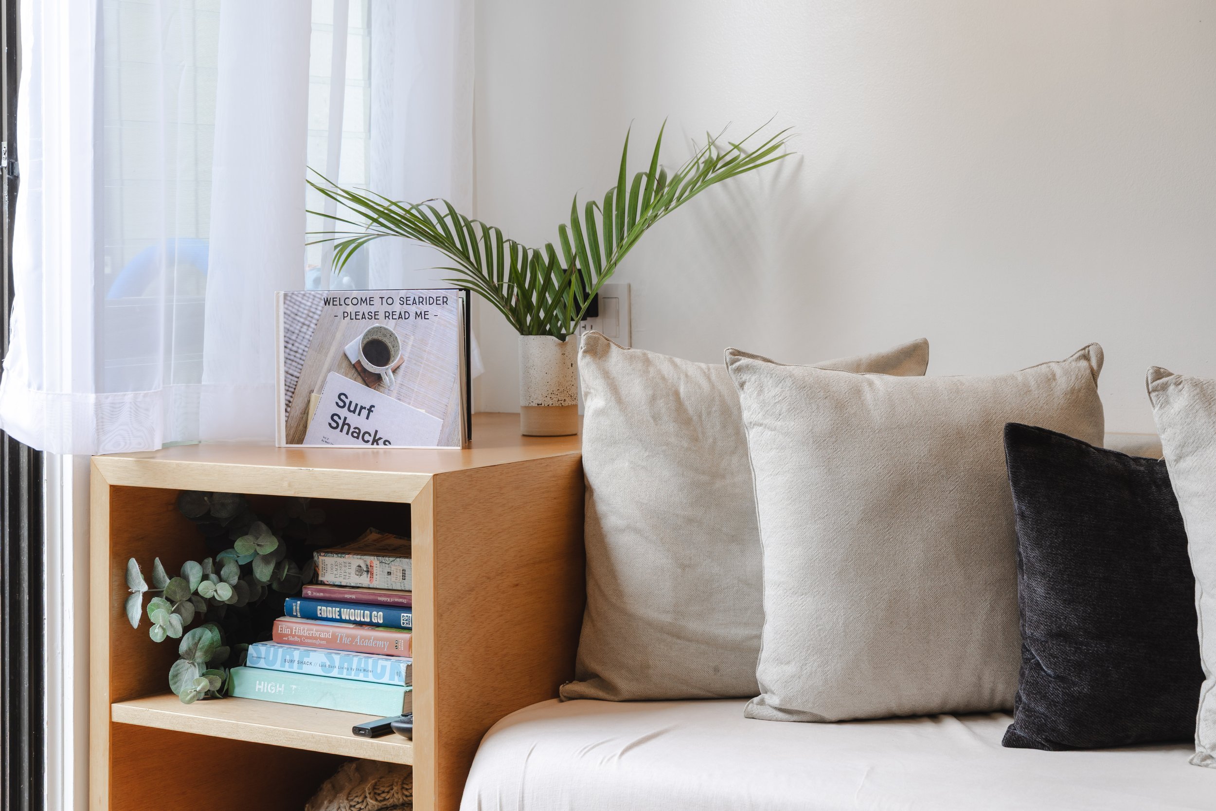 A cozy seating corner with beige cushions on a white sofa, a wooden side table with books about surfing and a small plant, a large green potted plant on the table, a framed sign reading 'Welcome to Seardier - Please Read Me', and sheer white curtains