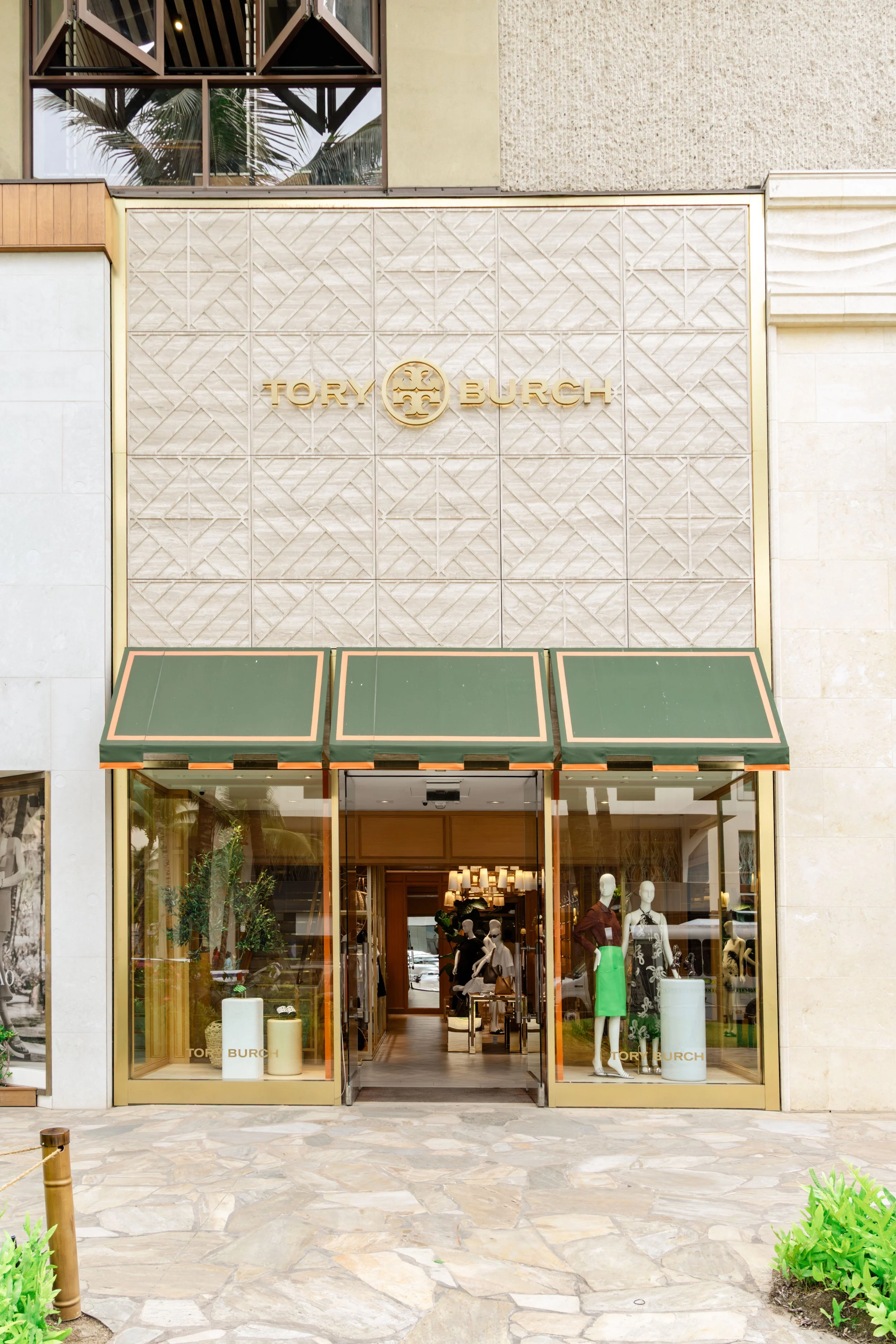 Storefront of Tory Burch with large glass windows displaying mannequins dressed in clothing, and the Tory Burch logo on the wall above the entrance.