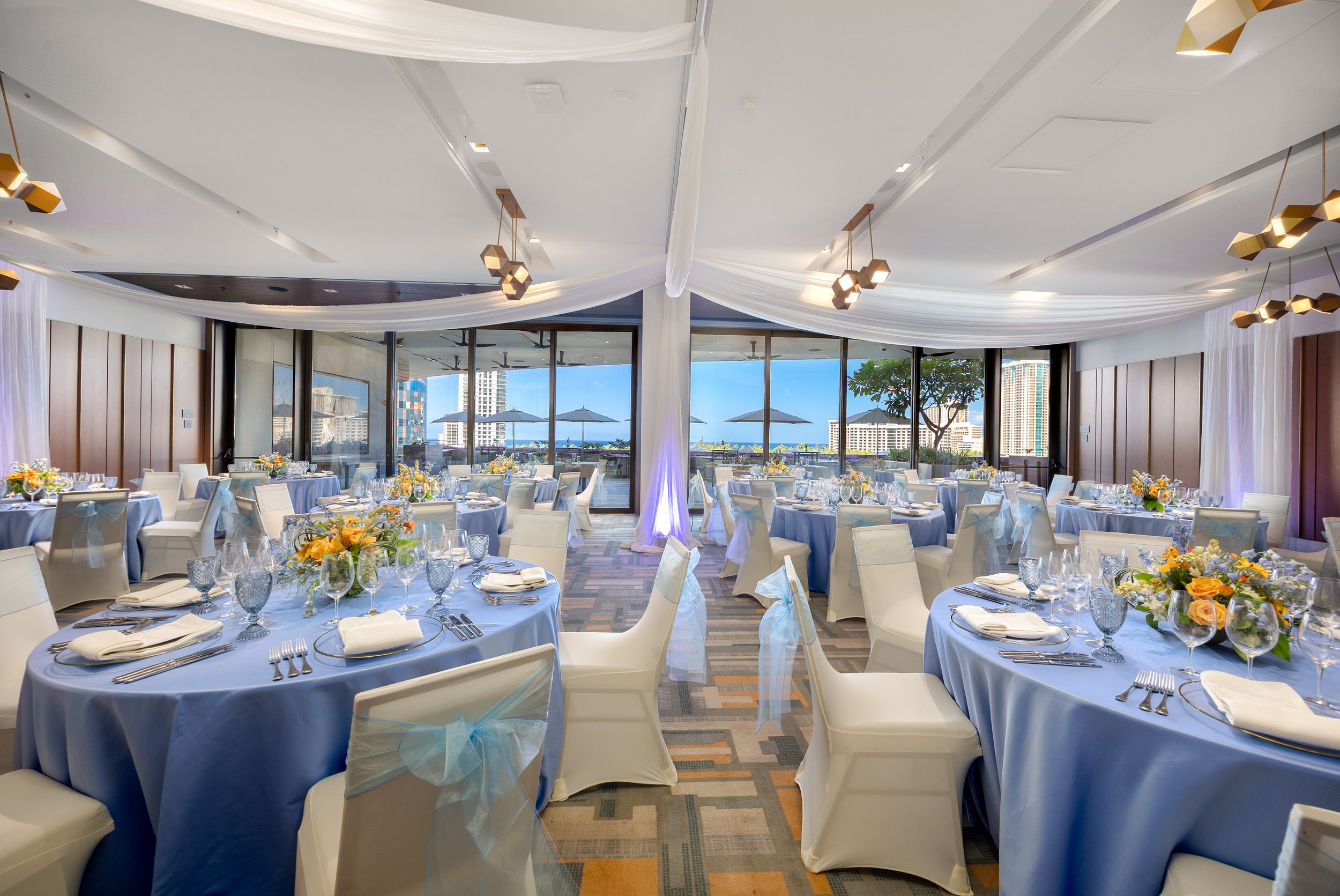 Elegant banquet hall decorated with round tables covered in light blue tablecloths, each set with white plates, silverware, glassware, and floral centerpieces. White chairs with light blue sashes surround the tables. The room has large windows with a
