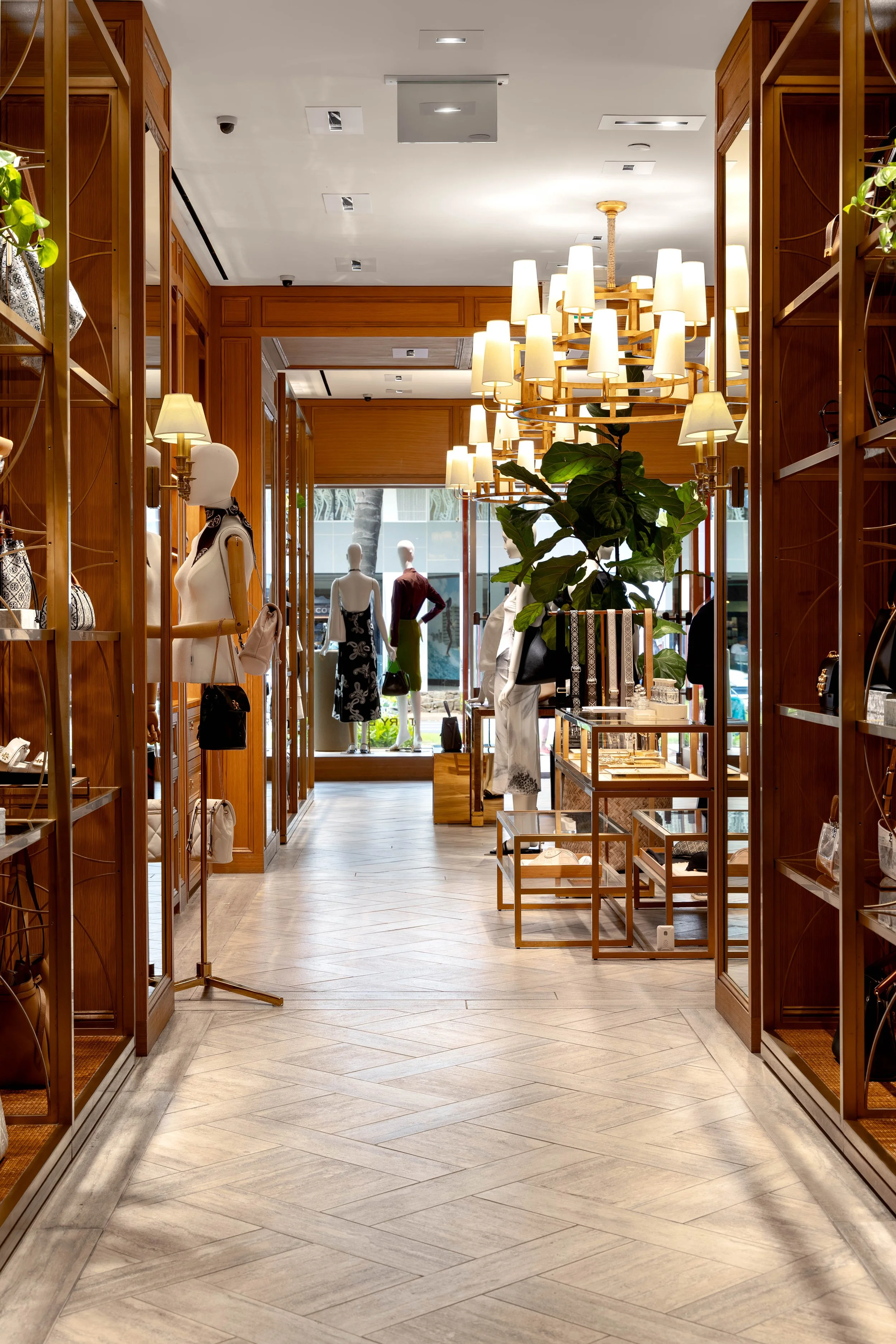 Interior of a retail store with wooden shelves displaying handbags, mannequins dressed in fashion, hanging plants, and a large chandelier with multiple lights.