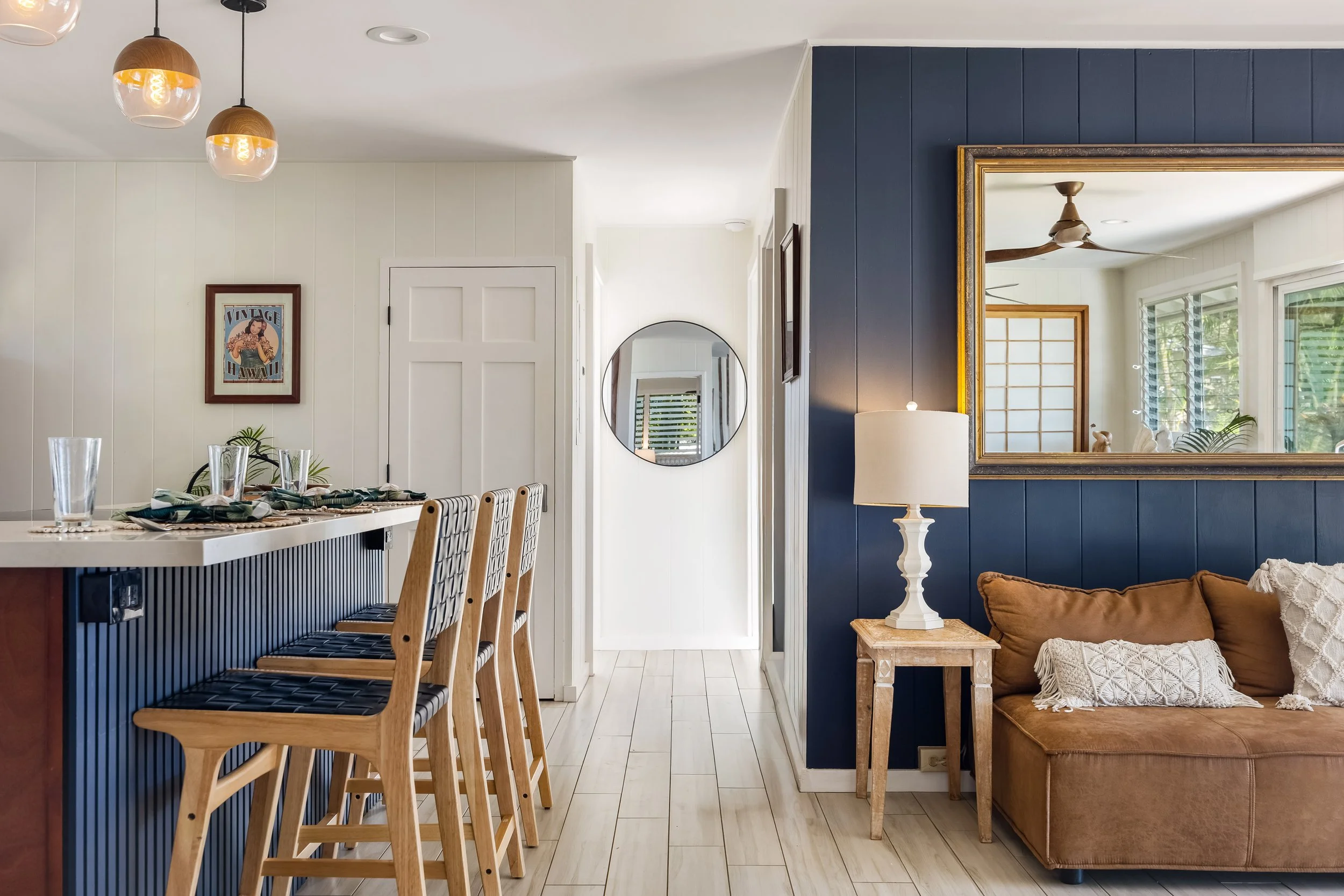 A modern living room with a dining area. The space features white and navy blue walls, a round mirror, and a vintage-style mirror frame. There's a brown sofa with a throw pillow, a wooden side table with a white lamp, and multiple windows with greene