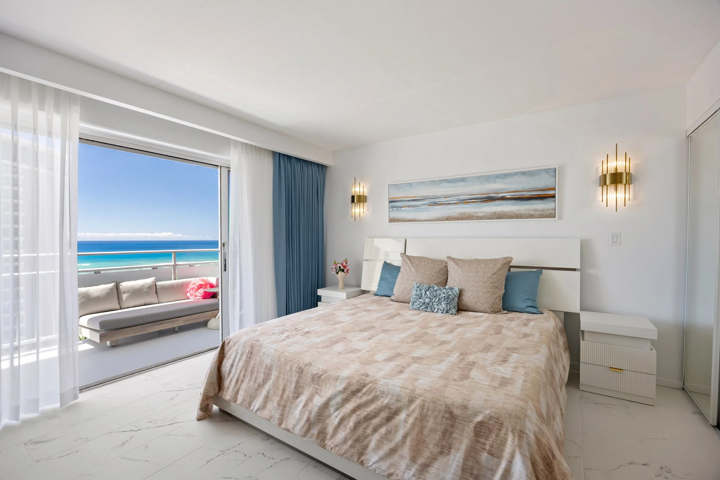 A modern bedroom with a view of the ocean through sliding glass doors. The room has white walls, a beige bedspread, and decorative pillows. There are wall-mounted golden light fixtures and a large abstract painting above the bed. A small white bedsid