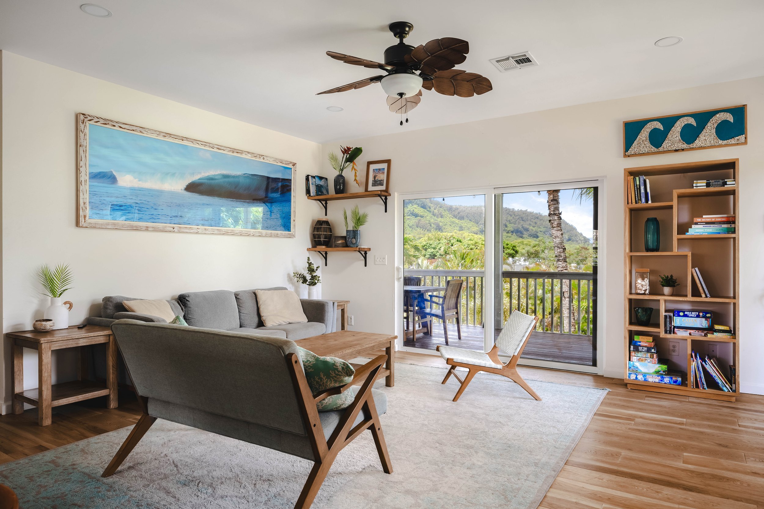 Living room with beige walls, large tropical landscape painting, wooden shelves with plants and books, window with view of greenery and mountains, sliding glass door leading to balcony with outdoor furniture, ceiling fan with leaf-shaped blades, gray
