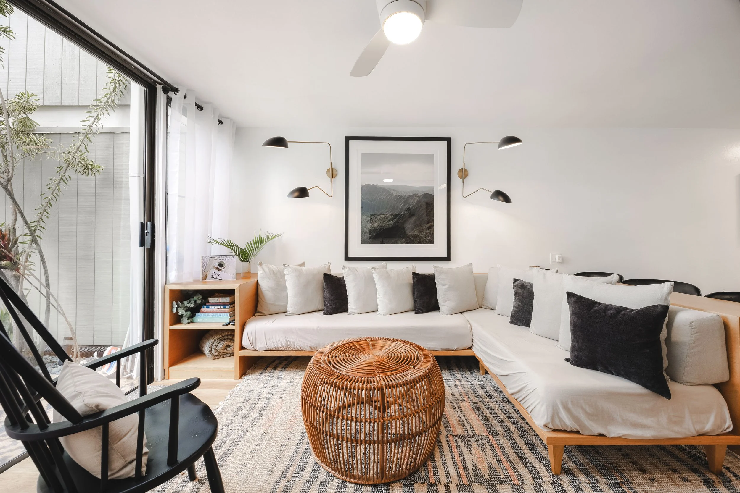 Living room with white sofa, black chairs, wicker coffee table, framed mountain artwork on white wall, wall-mounted black and gold lamps, sliding glass door with sheer white curtains, potted plant, and bookshelf.