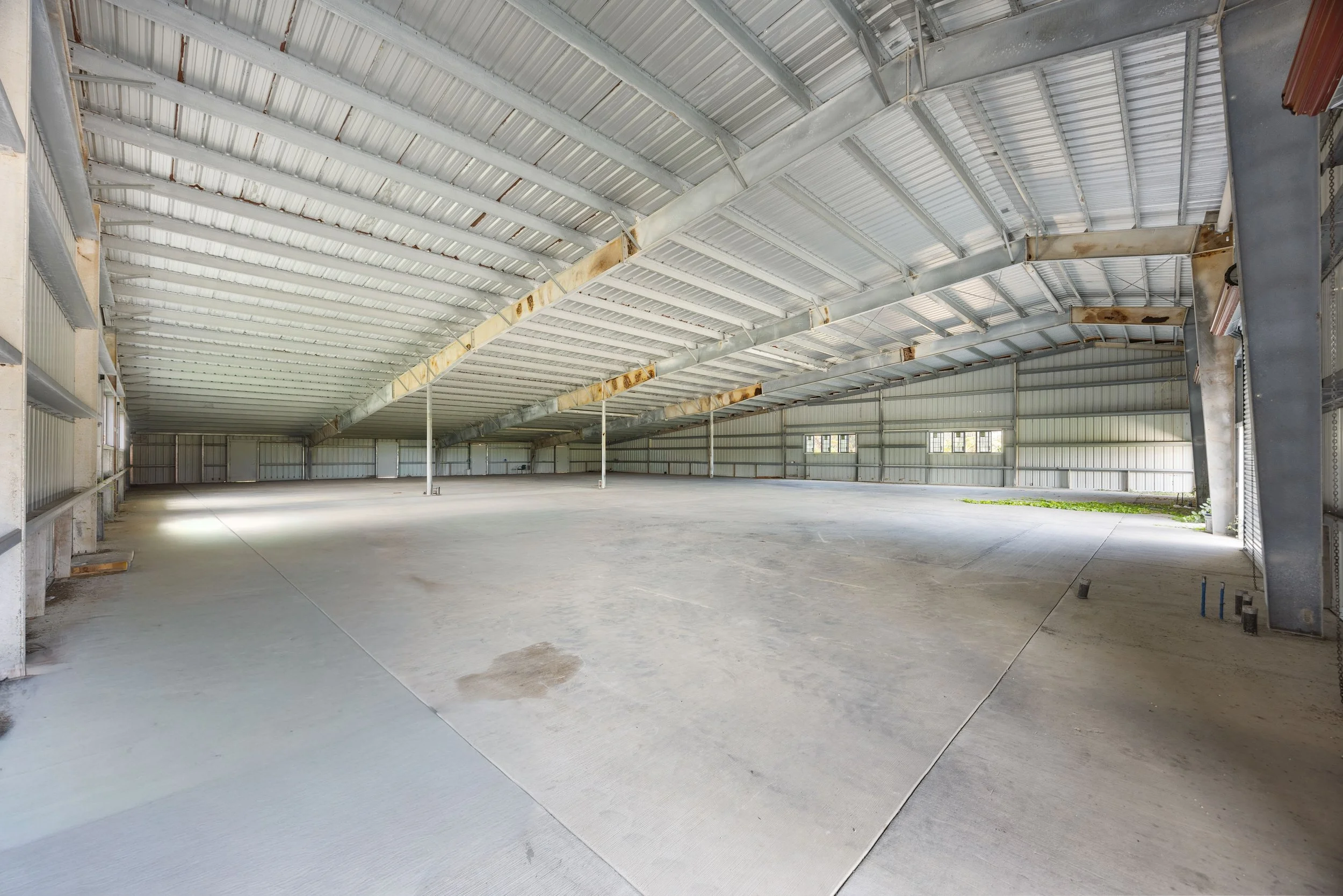 Empty large metal warehouse with concrete floor and metal roof and walls, sunlight coming through small windows.