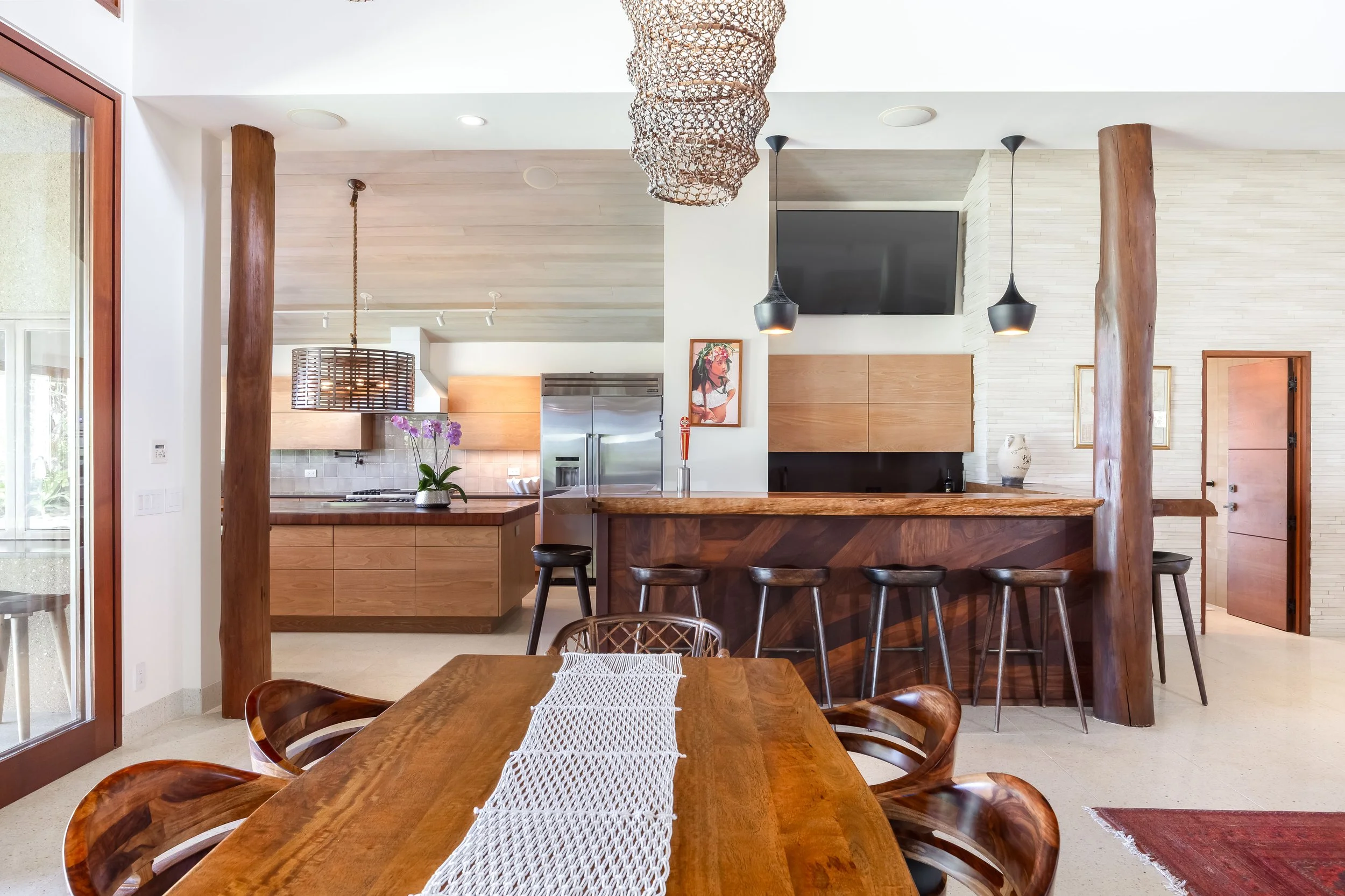 Living room with wooden furniture, a dining table with a white cloth, kitchen area in the background, decorative hanging lights, and large wooden support beams.