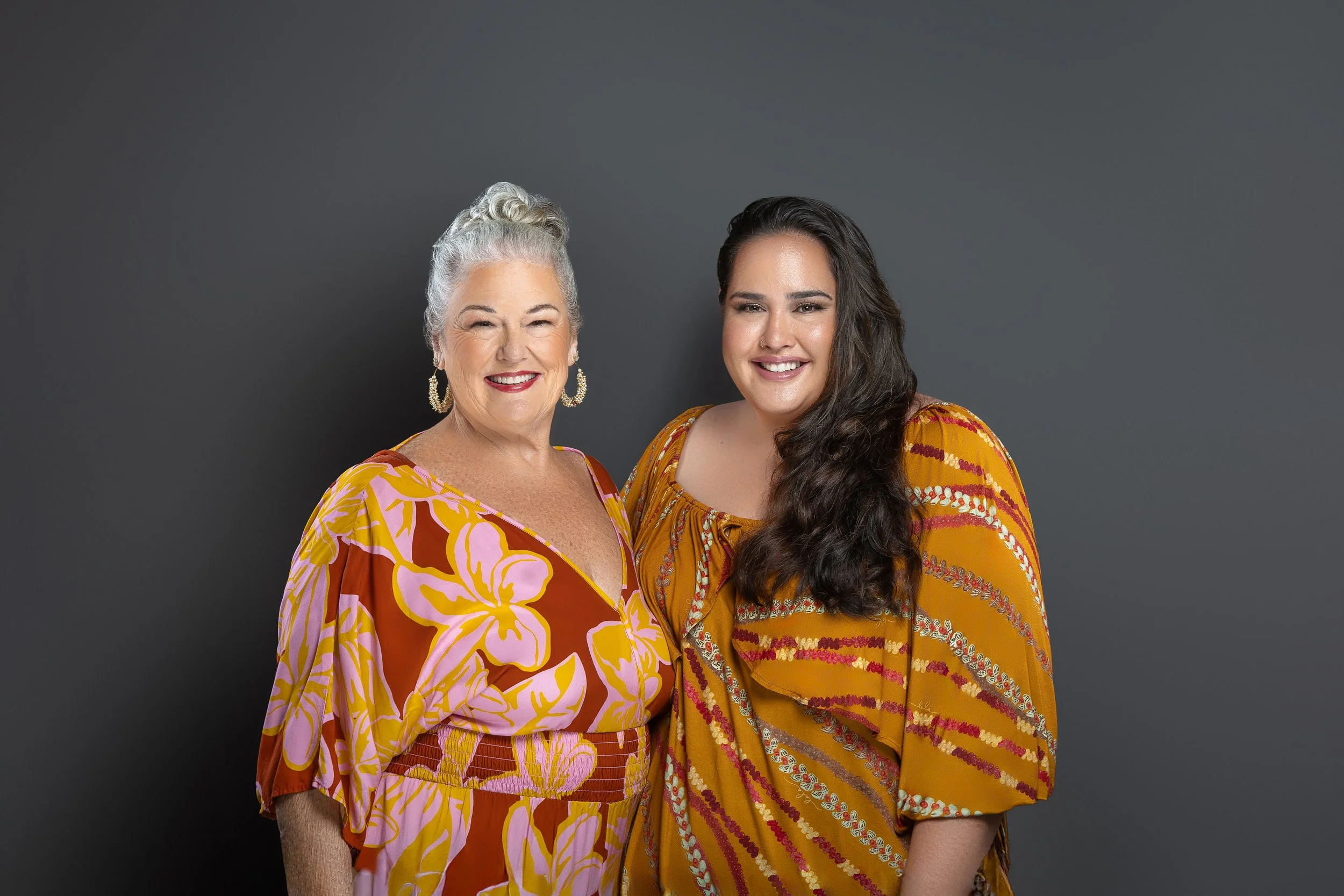Two women standing side by side and smiling against a gray background. The older woman has white hair styled in an updo, wearing large earrings and a brightly colored floral dress. The younger woman has long dark hair, wearing a patterned yellow dres