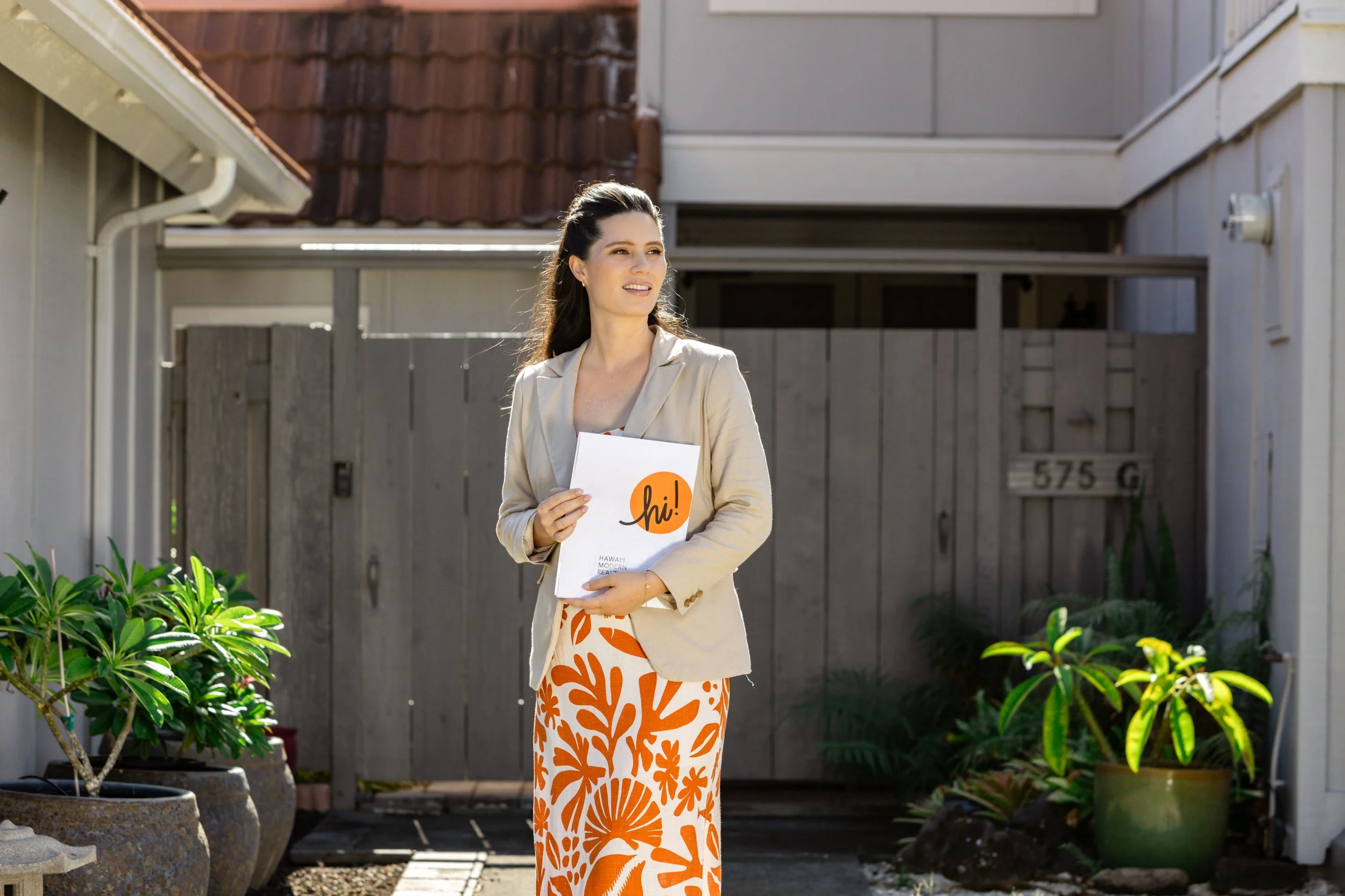 A woman in a beige blazer and orange floral pants standing outdoors, holding a white folder with an orange speech bubble and the word 'hi!'. She is smiling and looking to her left, with a gray wooden fence and potted plants in the background.