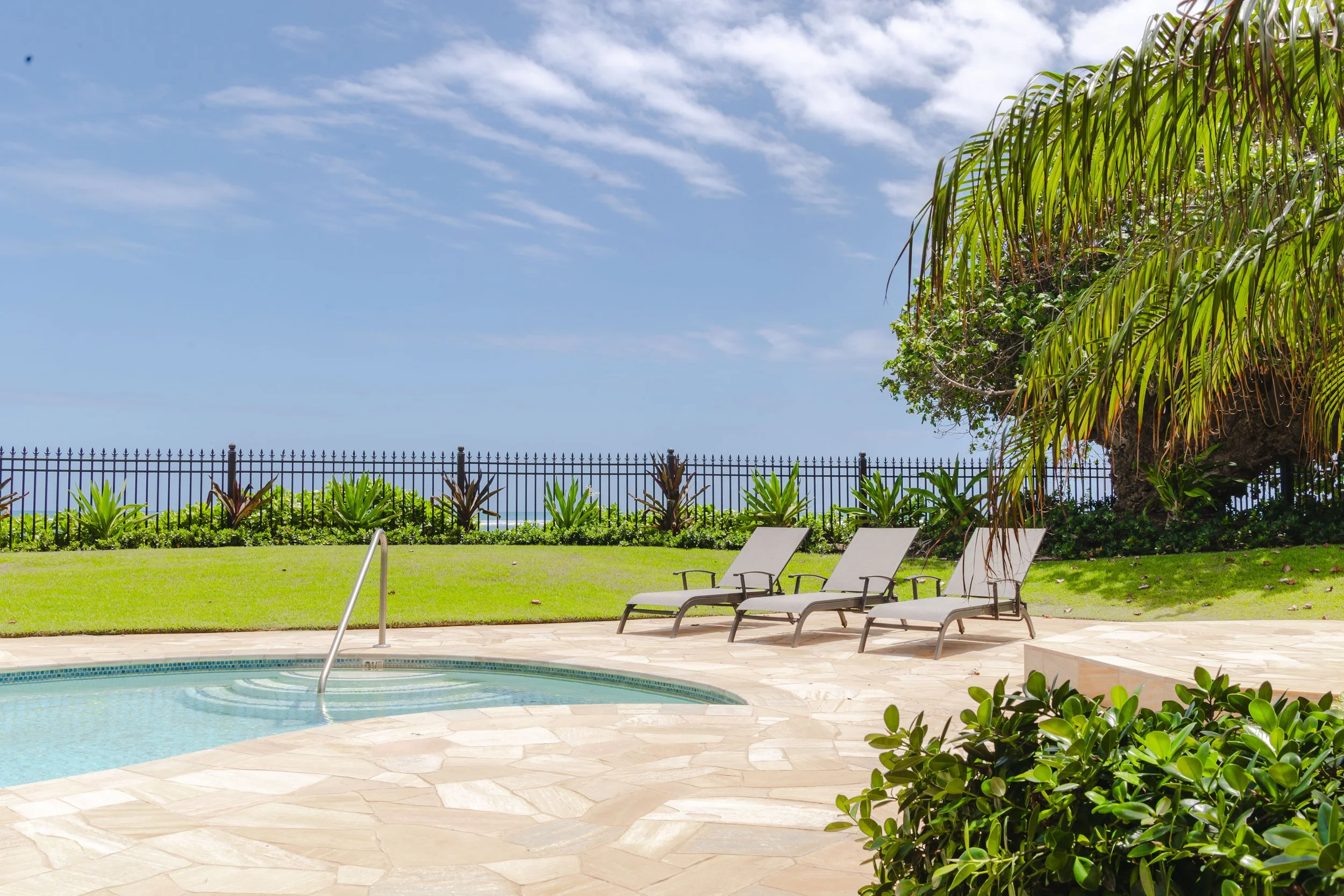 Swimming pool area with lounge chairs, lush green plants, a black metal fence, and a blue sky with clouds.