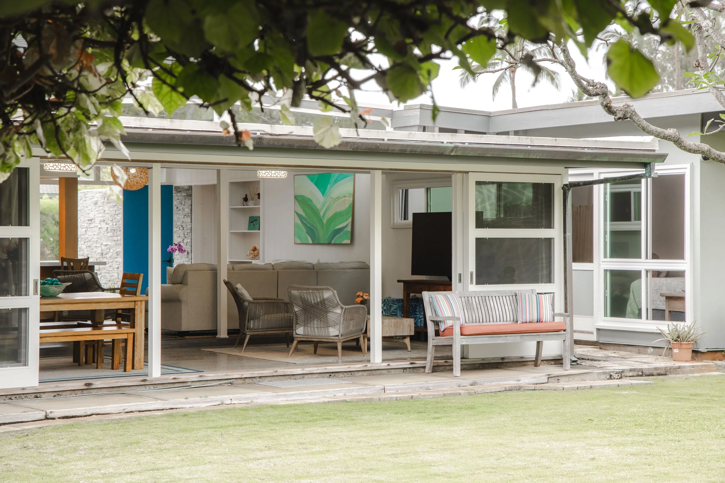 View of a house with an open patio, visible indoor living room with furniture and a painting, landscaped yard with green grass, and trees in the background.