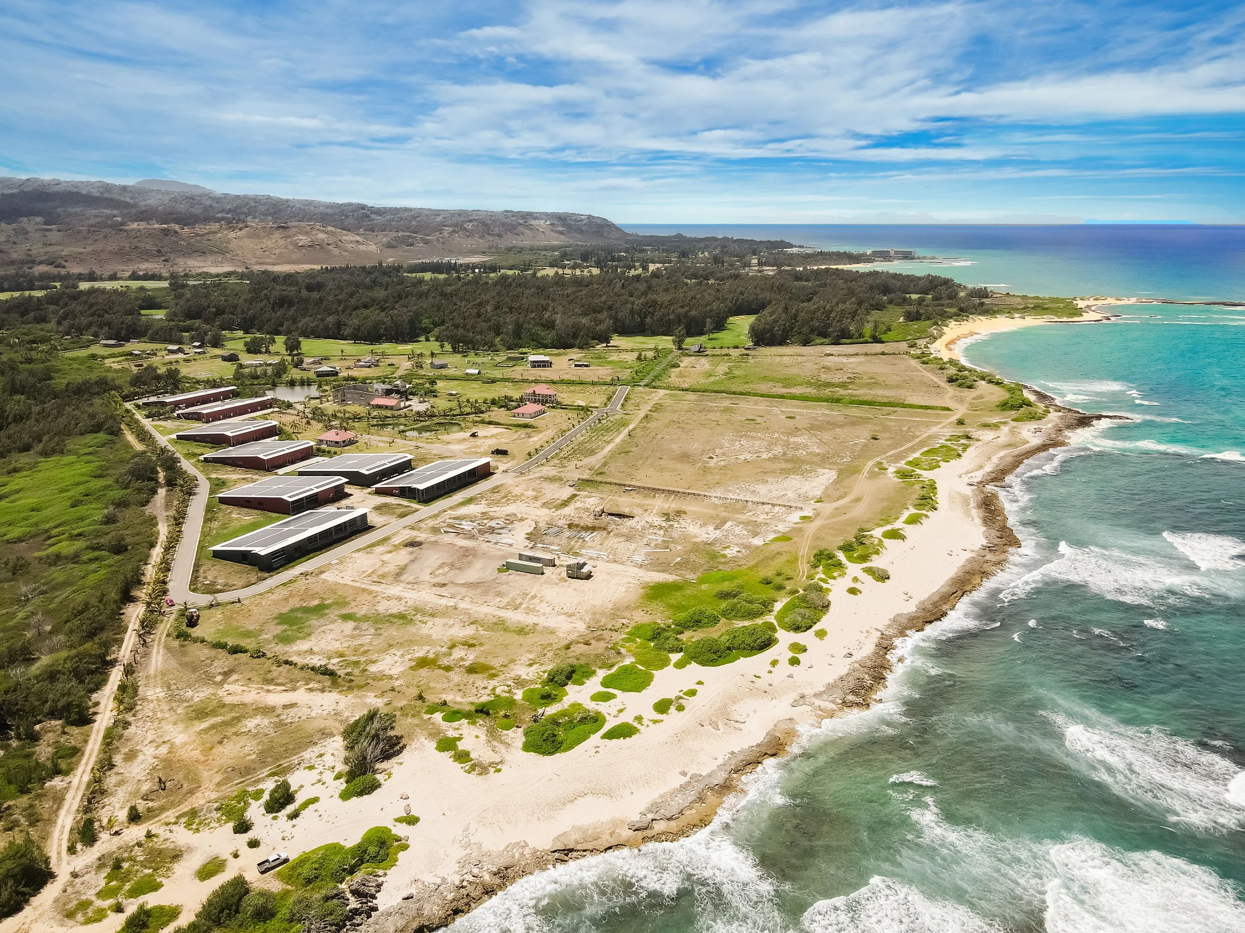 Aerial view of a coastal landscape showing a beach, ocean, green land, and mountains under a blue sky with clouds.