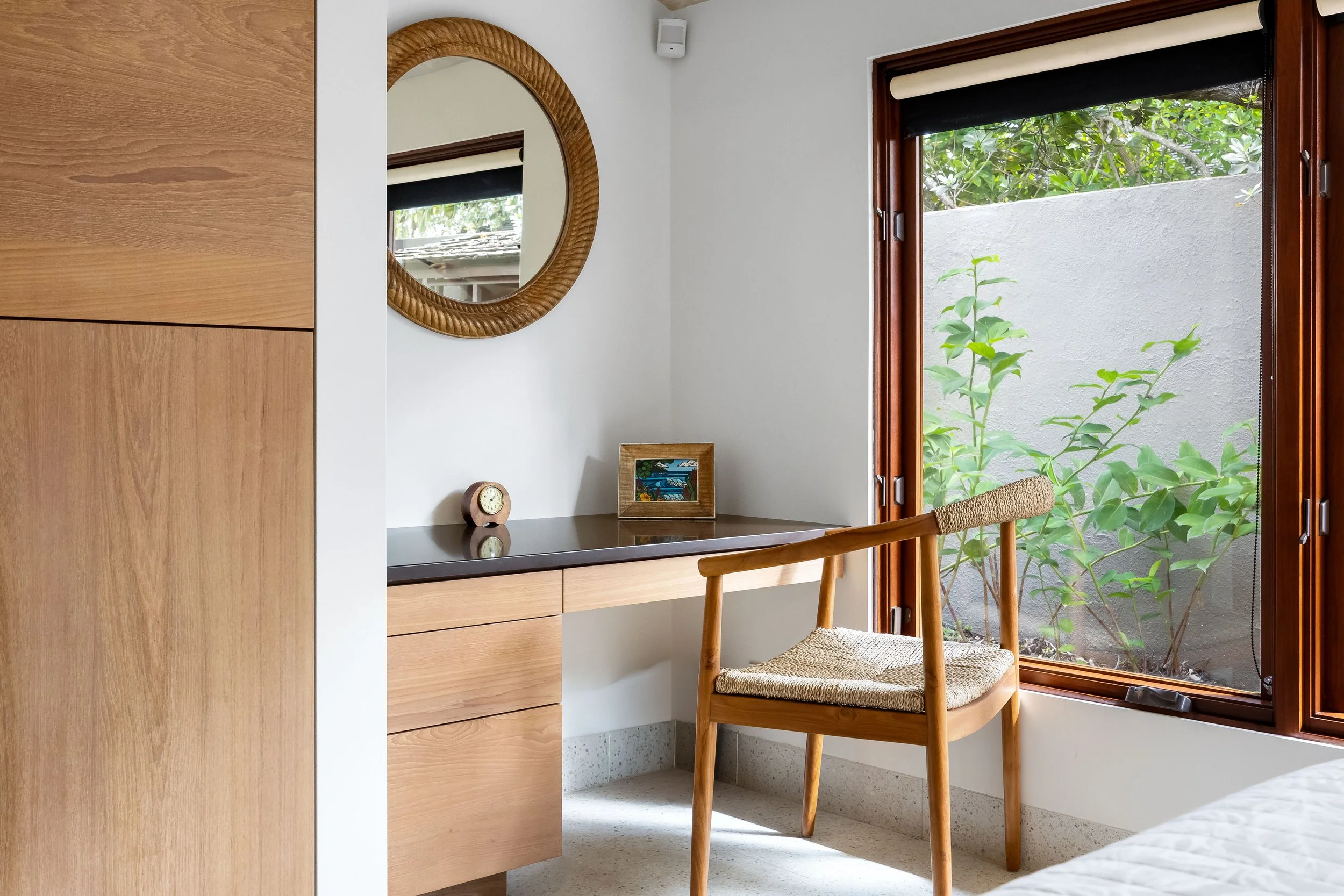 A cozy corner of a bedroom with a wooden chair with woven seat, a wooden dresser, a round mirror with a woven frame, a small clock, a small framed picture, a window with views of green foliage, and a part of a white bed in the foreground.
