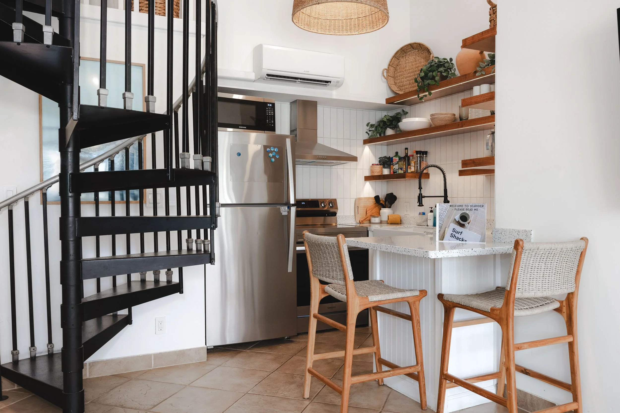 A kitchen with open wooden shelves holding bowls, plants, and jars. A stainless steel refrigerator, microwave, and oven are visible. A dining table with woven chairs and a black staircase are also part of the scene.