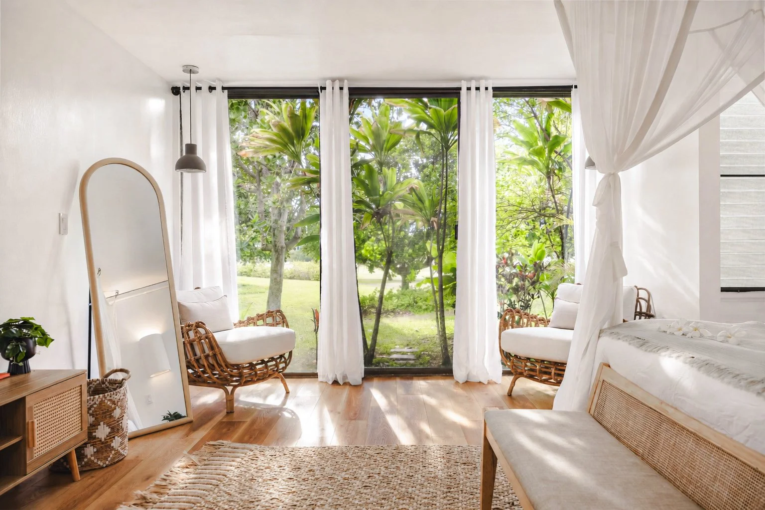 Bright bedroom with large sliding glass door overlooking a garden, white curtains, rattan chairs, a full-length mirror, a wooden dresser with plants, and a bed with white bedding