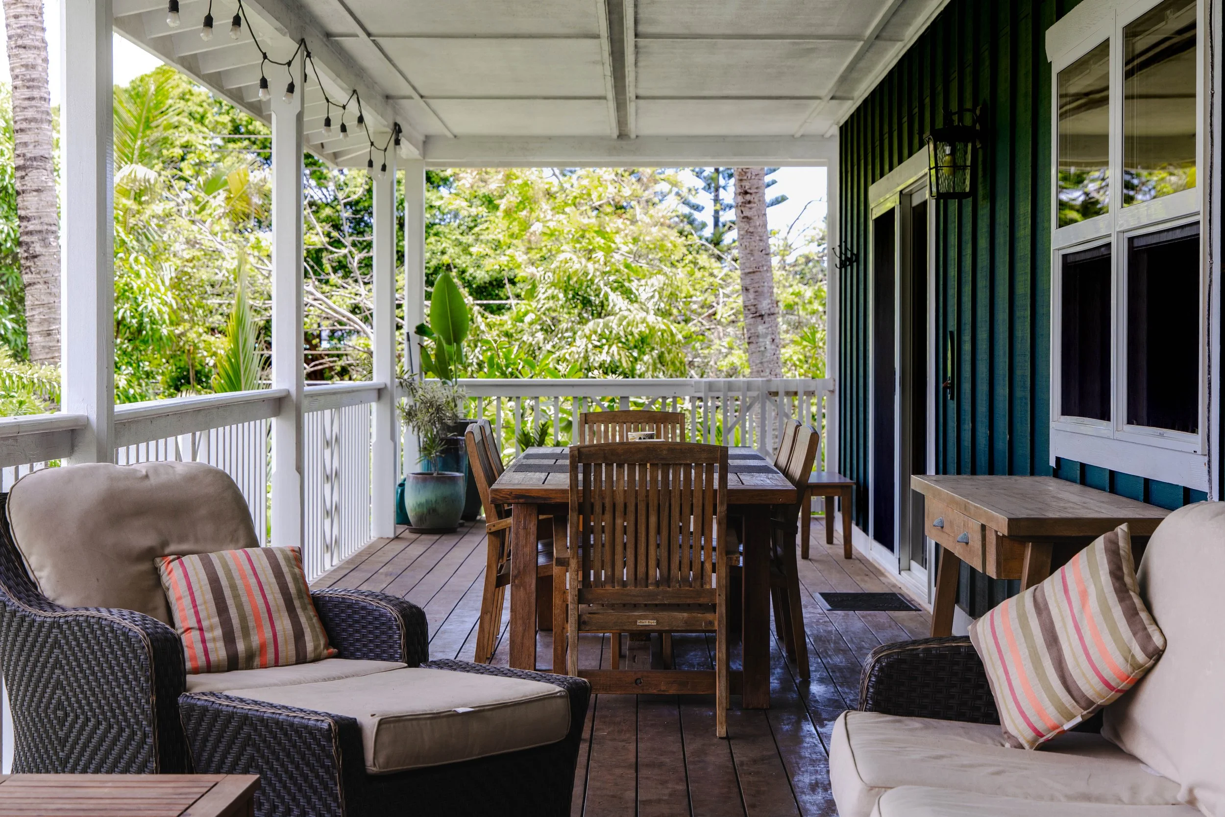 Covered porch with wooden dining table and chairs, wicker armchairs with cushions, potted plants, string lights, and lush greenery outside.