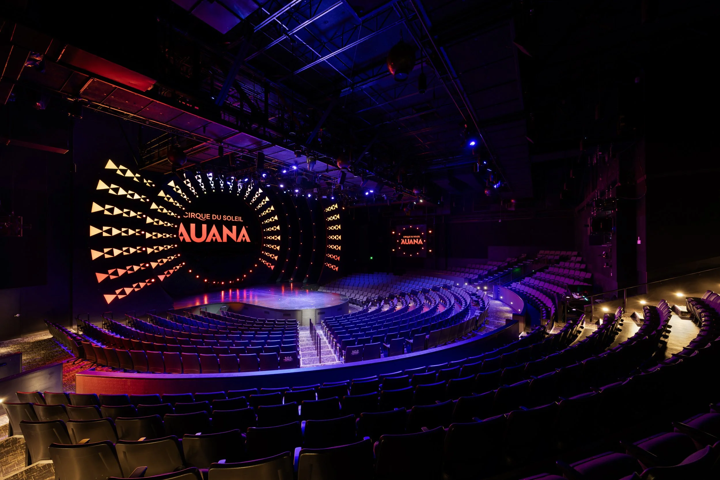 Inside view of a theater or performance venue with empty seats illuminated by colorful stage lights. There is a large circular screen displaying the words 'Cirque du Soleil AUANA' along with geometric designs.