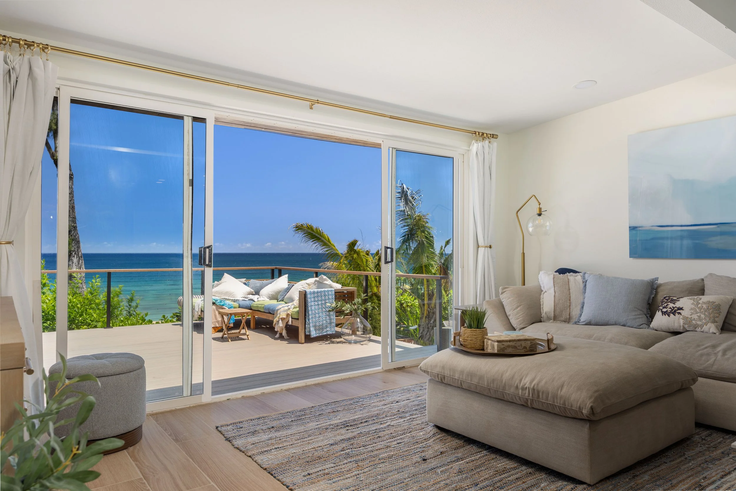 Living room with large glass sliding doors opening to a balcony with ocean view, white curtains, beige sectional sofa, side table with plants, and artwork on the wall.