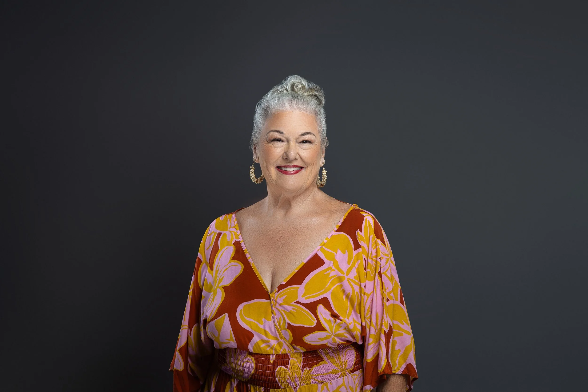 An elderly woman with grey hair styled in a proud updo, wearing a colorful, floral-patterned dress with pink, orange, and yellow hues, smiling confidently against a dark grey background.
