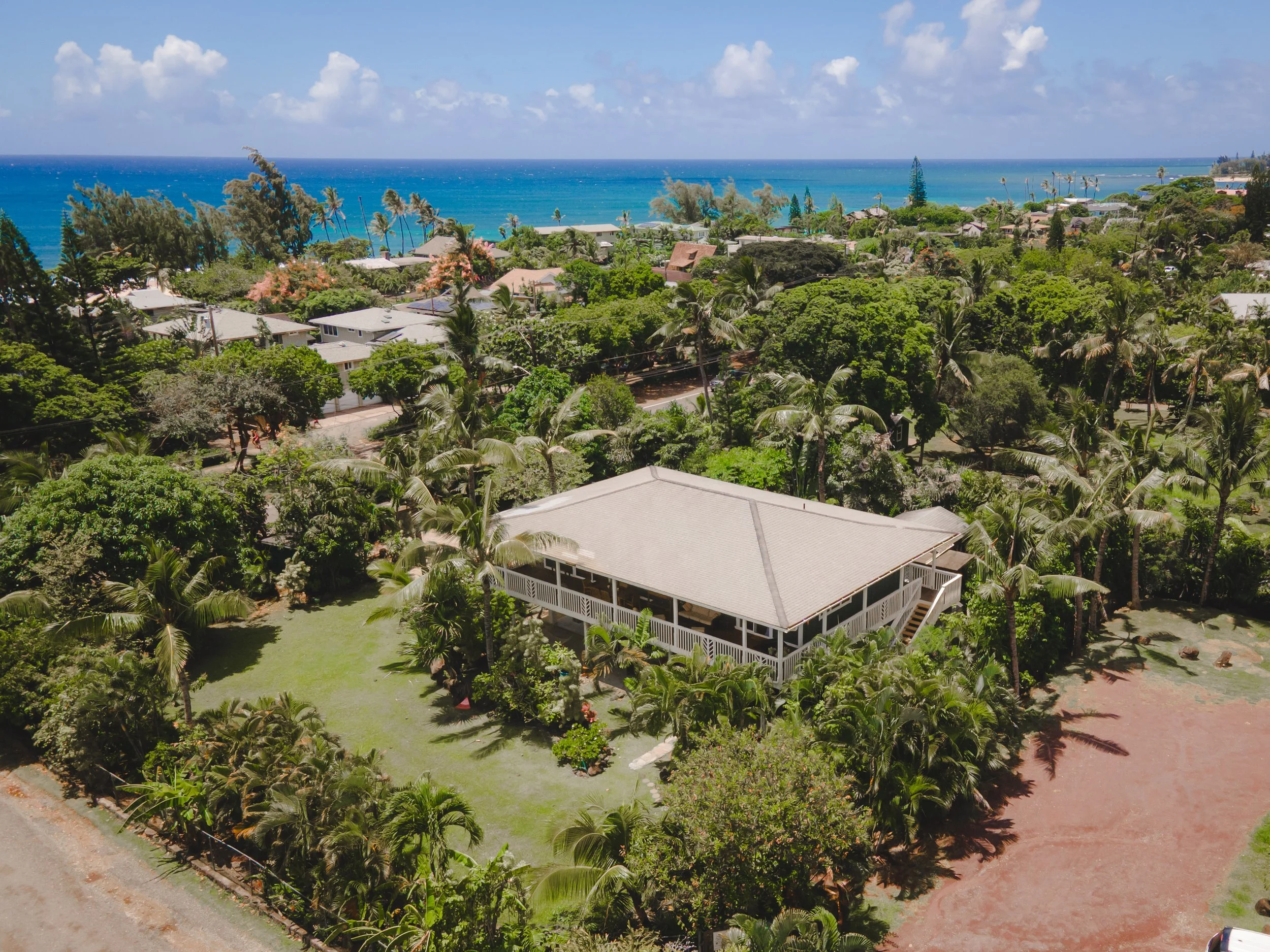 Aerial view of a tropical coastal neighborhood with lush greenery, houses, and a blue ocean in the background under a partly cloudy sky.
