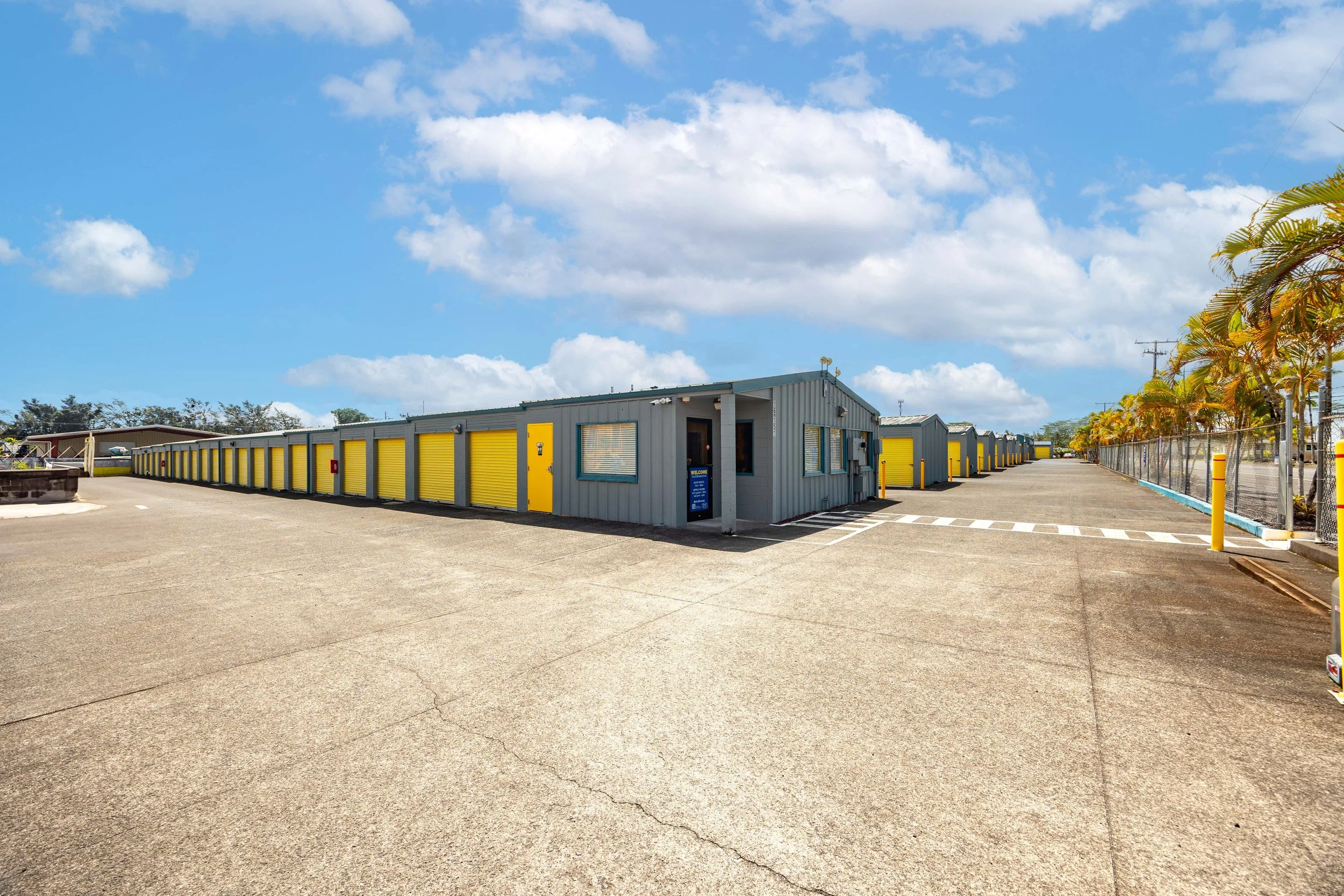 A row of storage units with bright yellow doors under a partly cloudy blue sky, with a paved parking area and some palm trees on the right side.