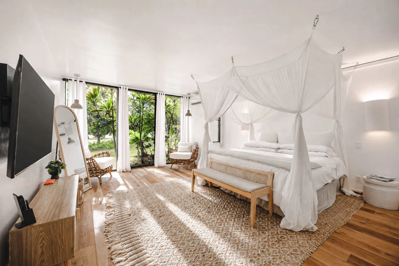 Bright bedroom with a white canopy bed, large sliding glass doors with white curtains, and views of greenery outside.