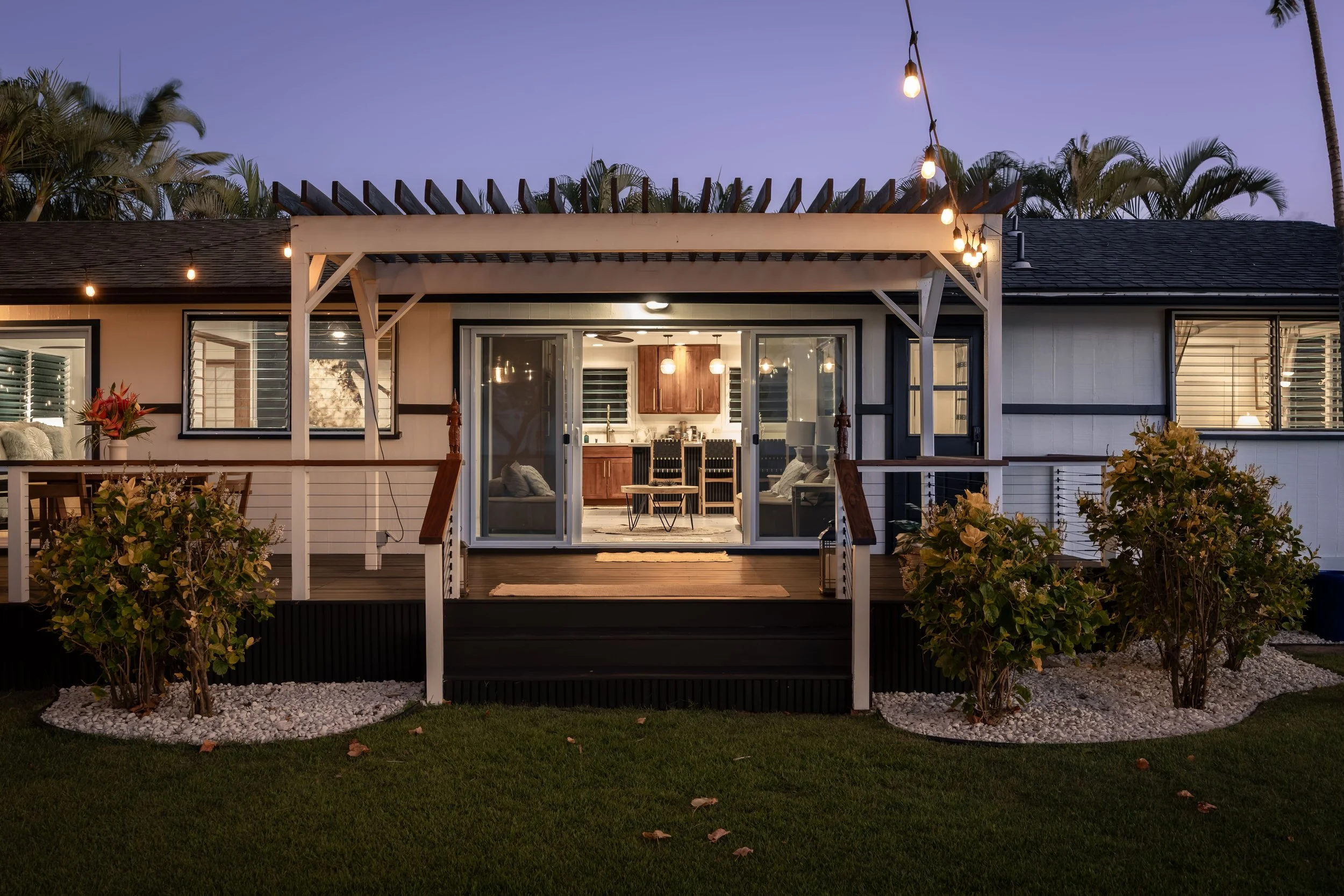 A cozy backyard patio with string lights hanging above and an open sliding door leading to an indoor kitchen and dining area, with lush greenery and flowers in the foreground at dusk.