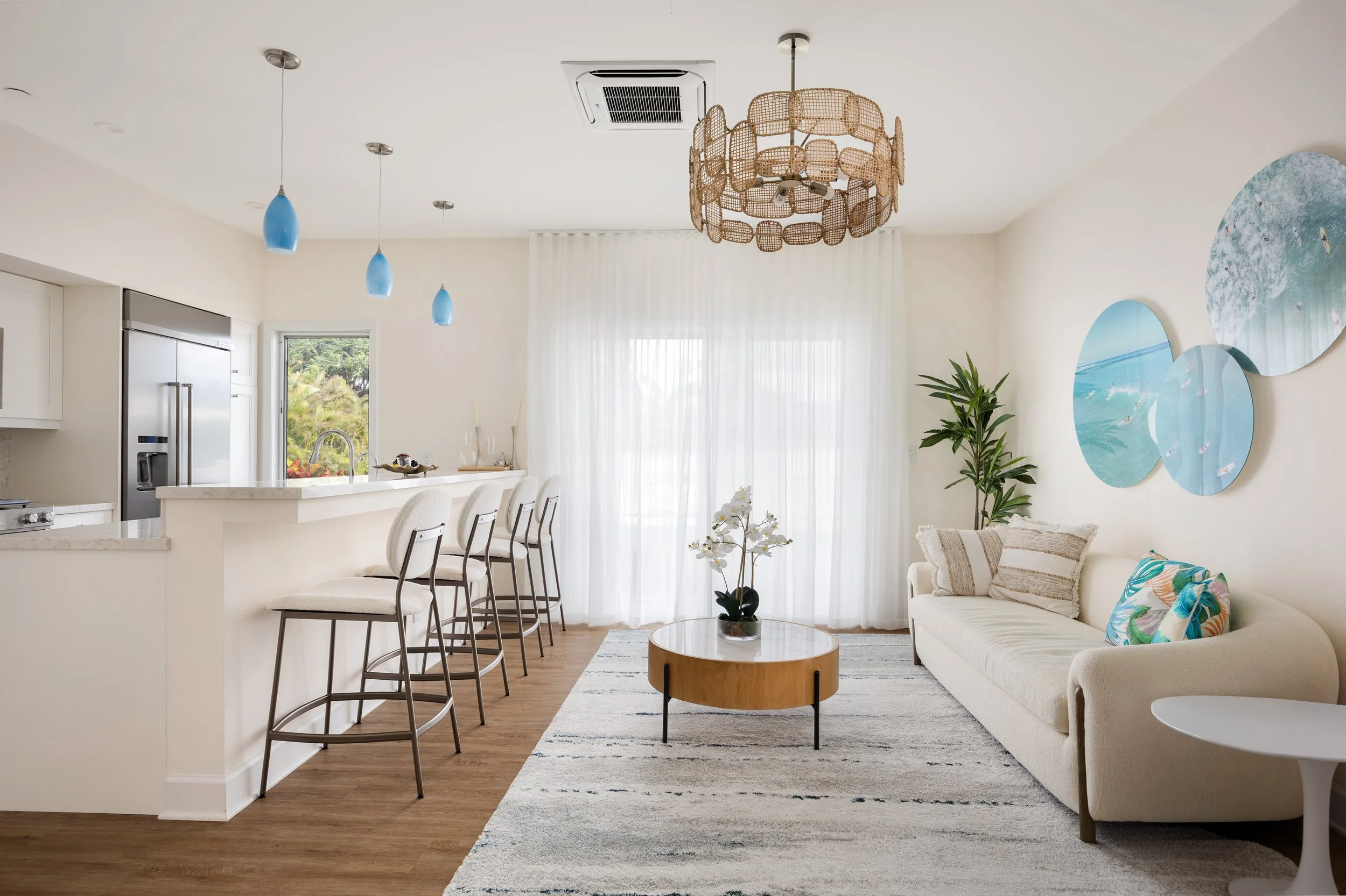 A bright living room with a white sofa decorated with colorful pillows, a round wooden coffee table, a potted orchid, and seaside-themed artwork on the wall, illuminated by natural light through sheer curtains.