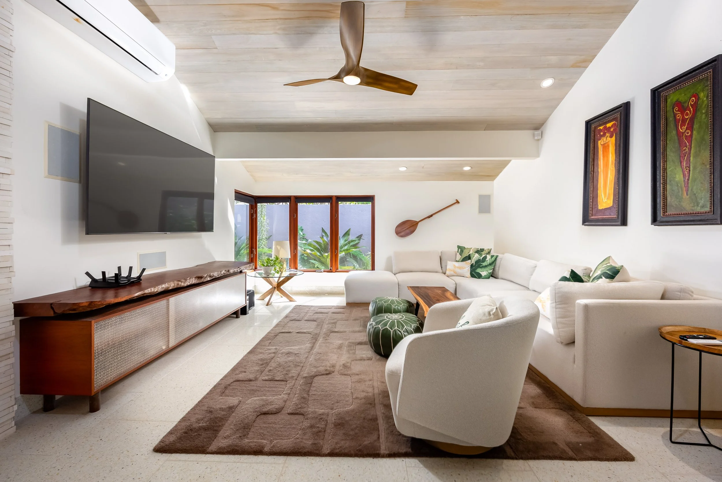 A bright living room with a large white sectional sofa, green patterned pillows, and two green leather poufs. A wooden coffee table is in front. On the wall, there are two framed colorful abstract paintings. A large flat screen TV hangs on the wall, 