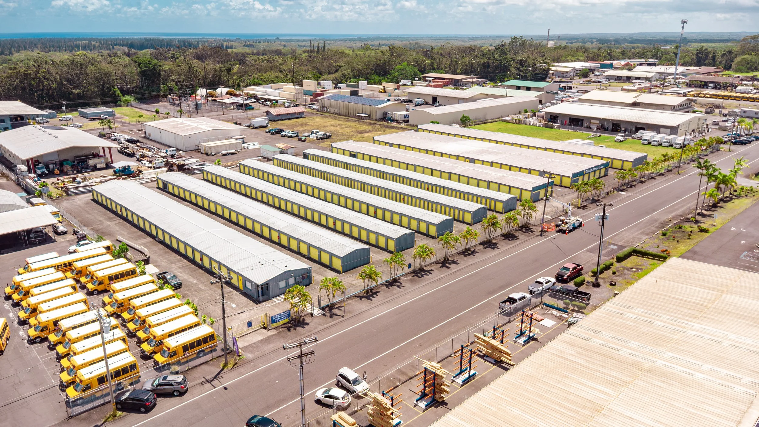 Aerial view of a storage facility with rows of yellow school buses, multiple industrial warehouses, parking lots, and green areas with trees, under a partly cloudy sky.