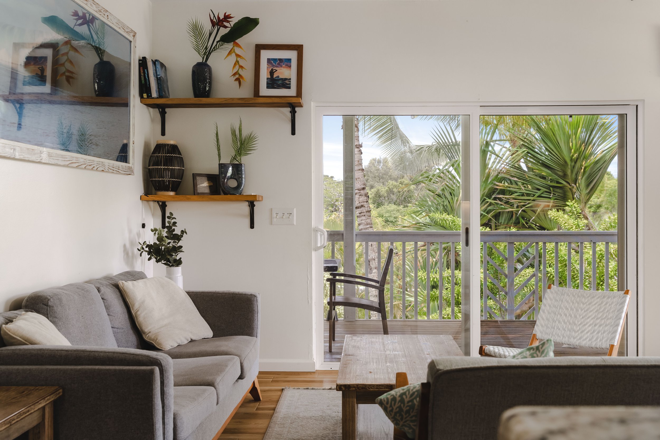Living room with gray sofa with cushions, wooden coffee table, chairs on a balcony, sliding glass door, and tropical plant scenery outside.