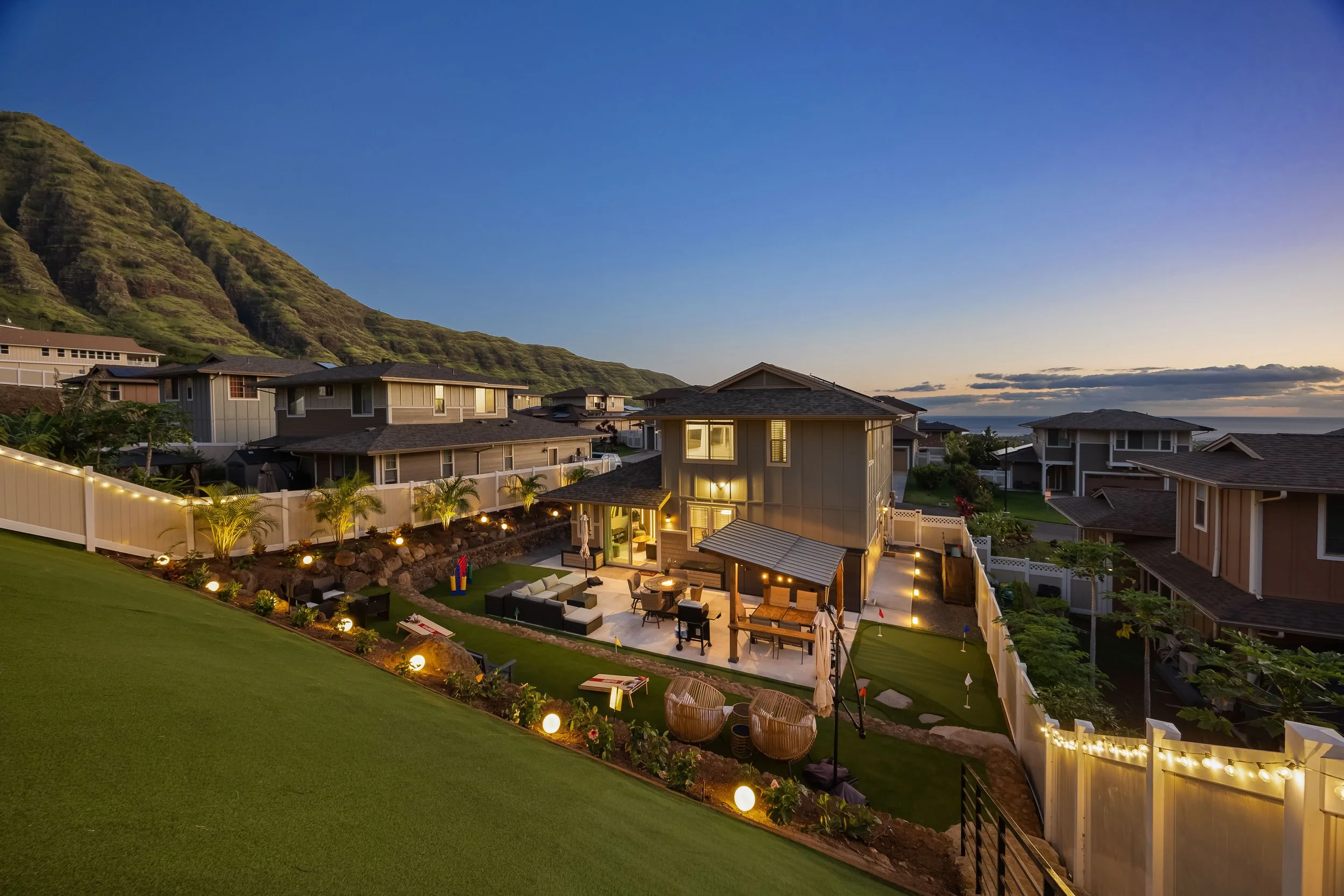 A residential backyard with outdoor seating, a small play area, and string lights, with neighboring houses and a green mountain in the background during dusk.