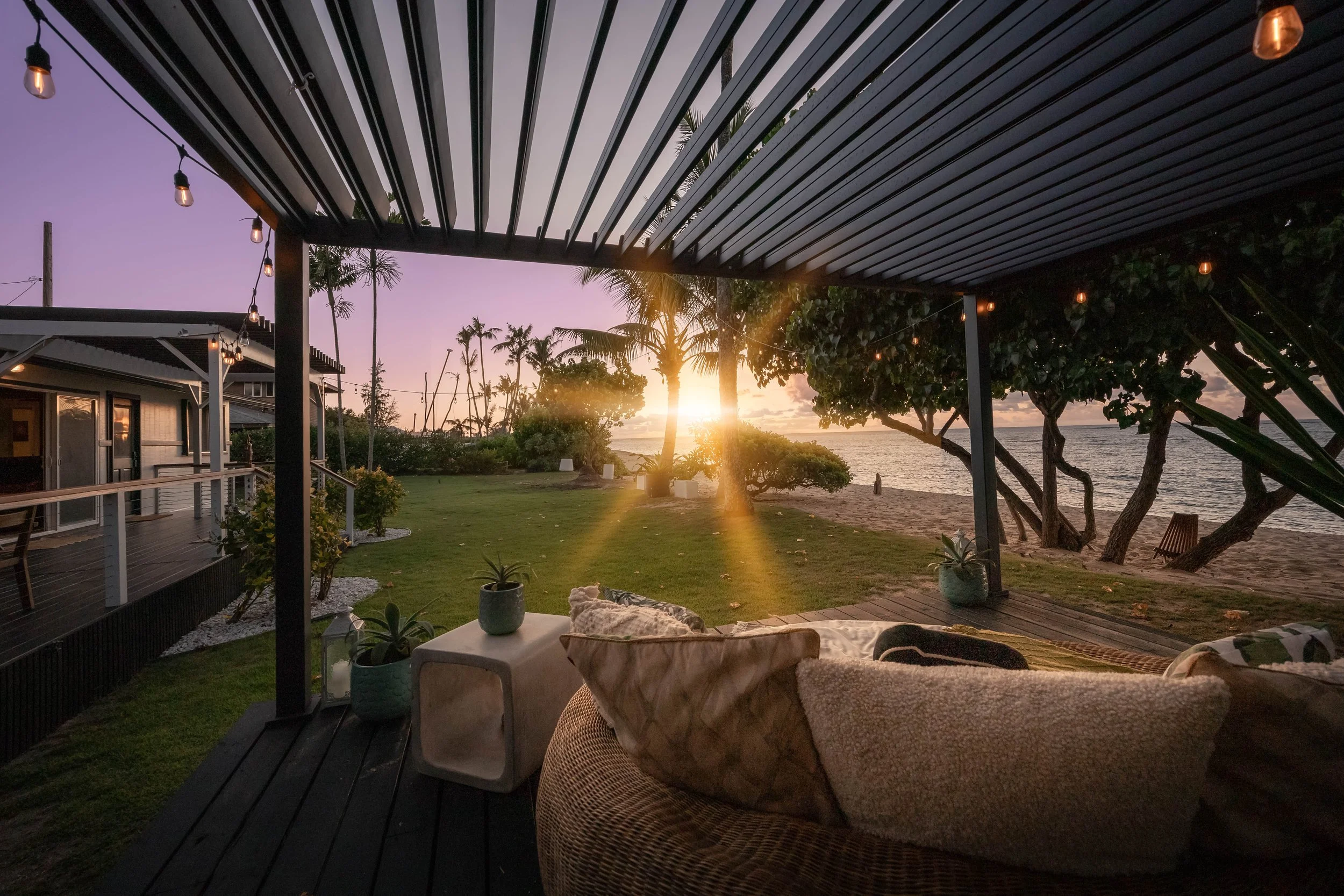 View from a covered outdoor patio overlooking a beach with palm trees at sunset, with hanging string lights and outdoor furniture.