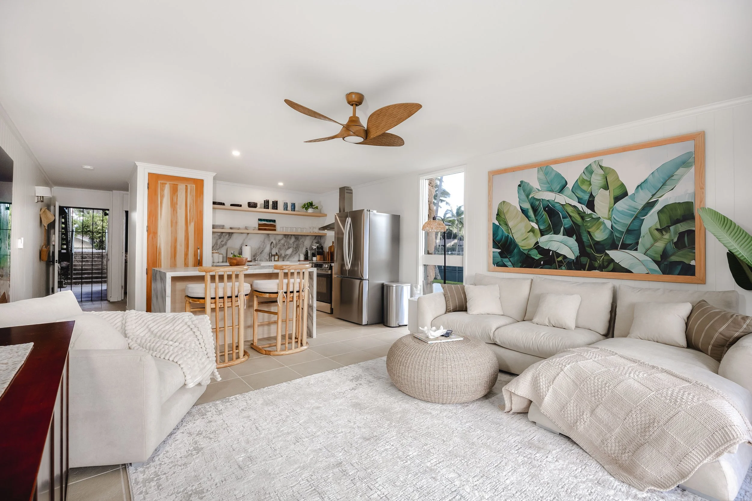 Living room with white sofas, a large tropical leaf painting, a woven pouf, and a kitchen area with bar stools and open shelves.