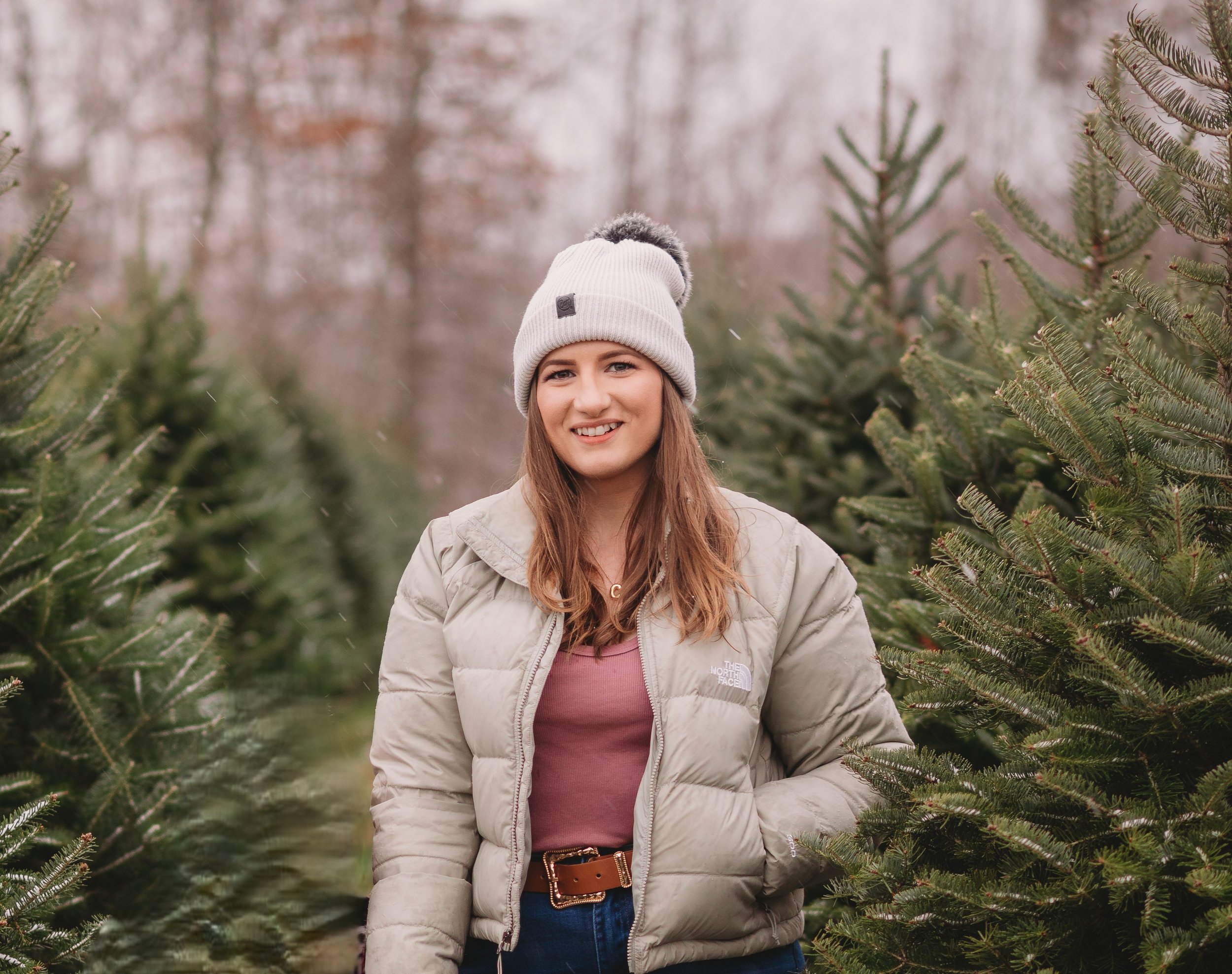 A young woman with long brown hair smiling outdoors between Christmas trees, wearing a beige puffer jacket, a pink top, a light gray beanie, and a brown belt.