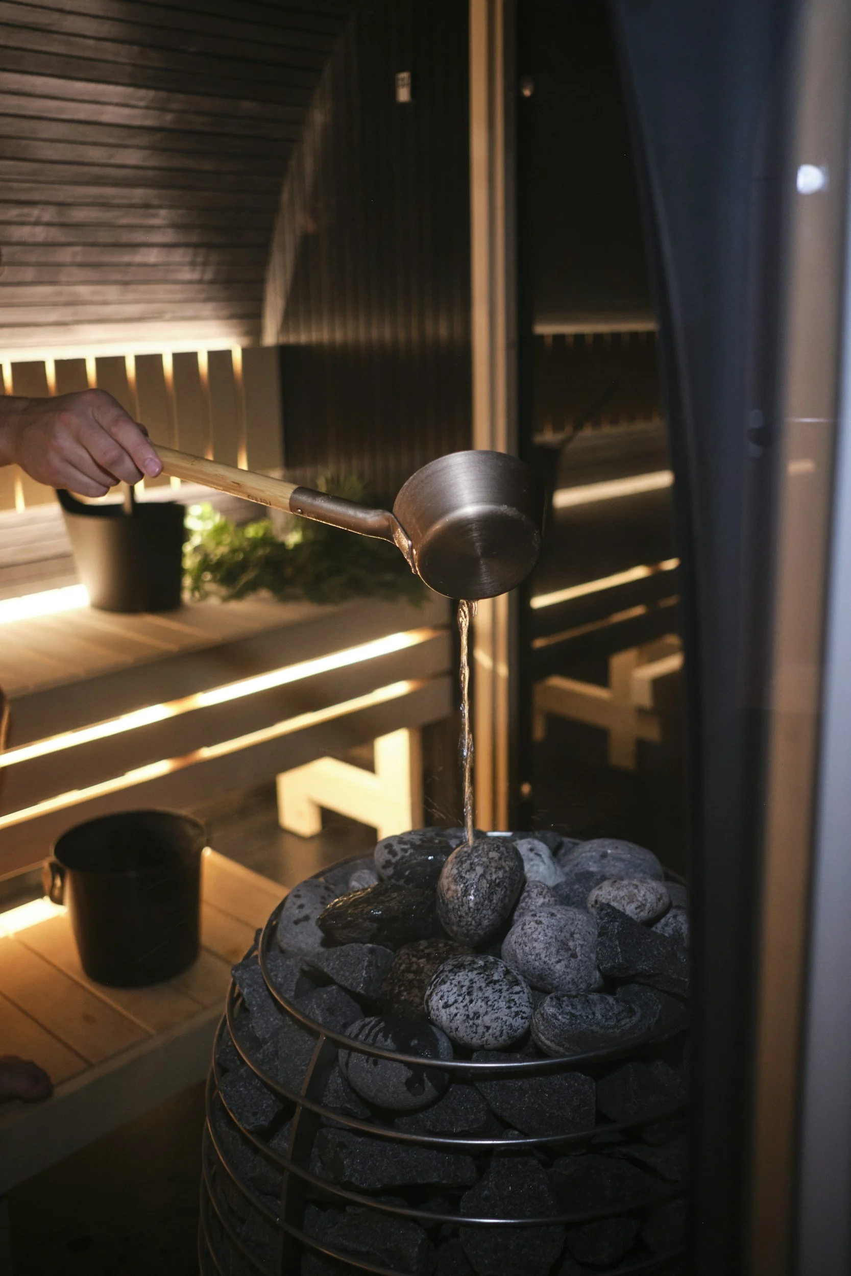 A hand holding a wooden ladle pouring water over rocks in a sauna stone heater, with wooden walls and benches visible in the background.
