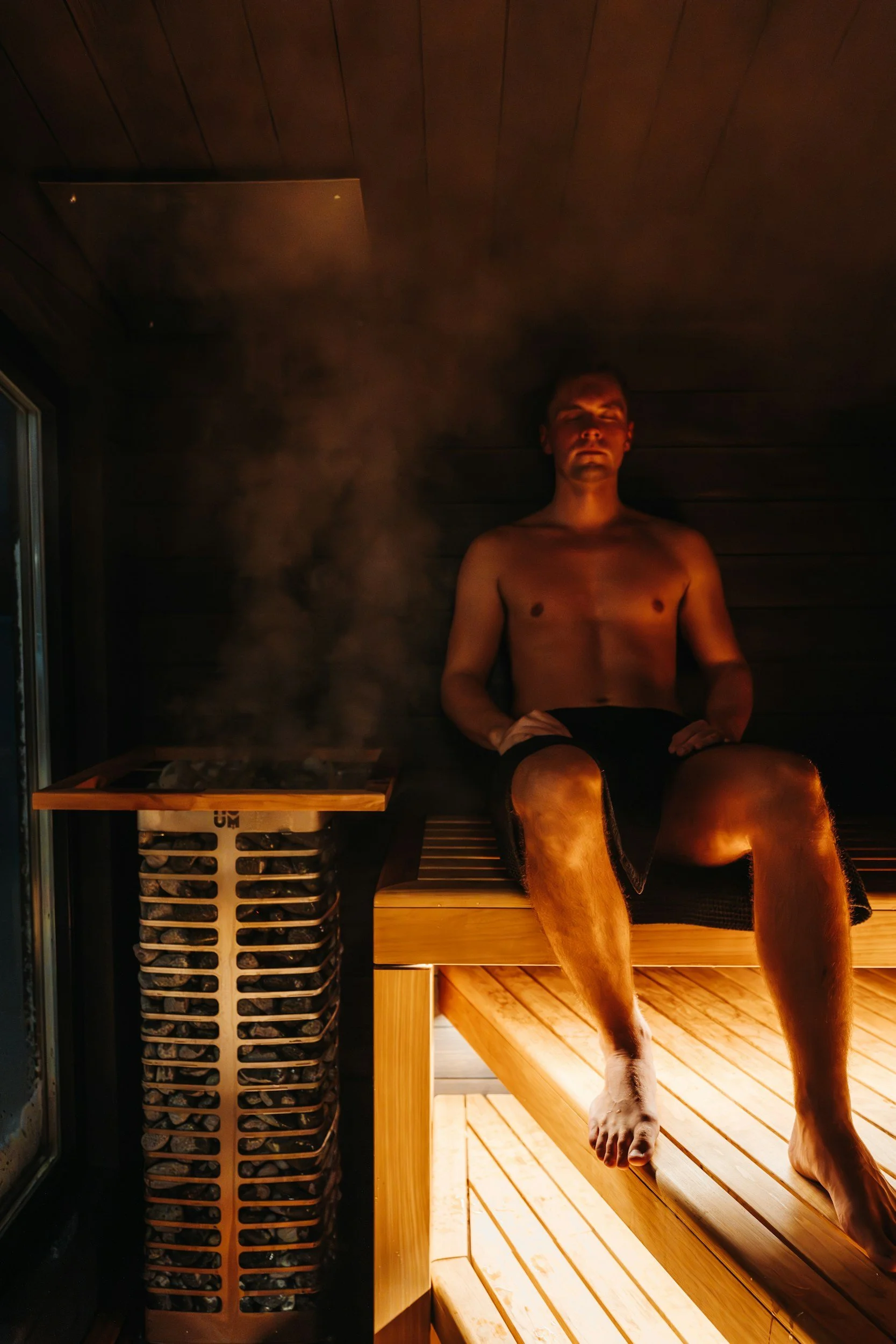 A man resting in a sauna sits on a wooden bench with his eyes closed. Steam is visible in the infrared sauna, and a pile of rocks is placed in a heater to generate steam inside the sauna.