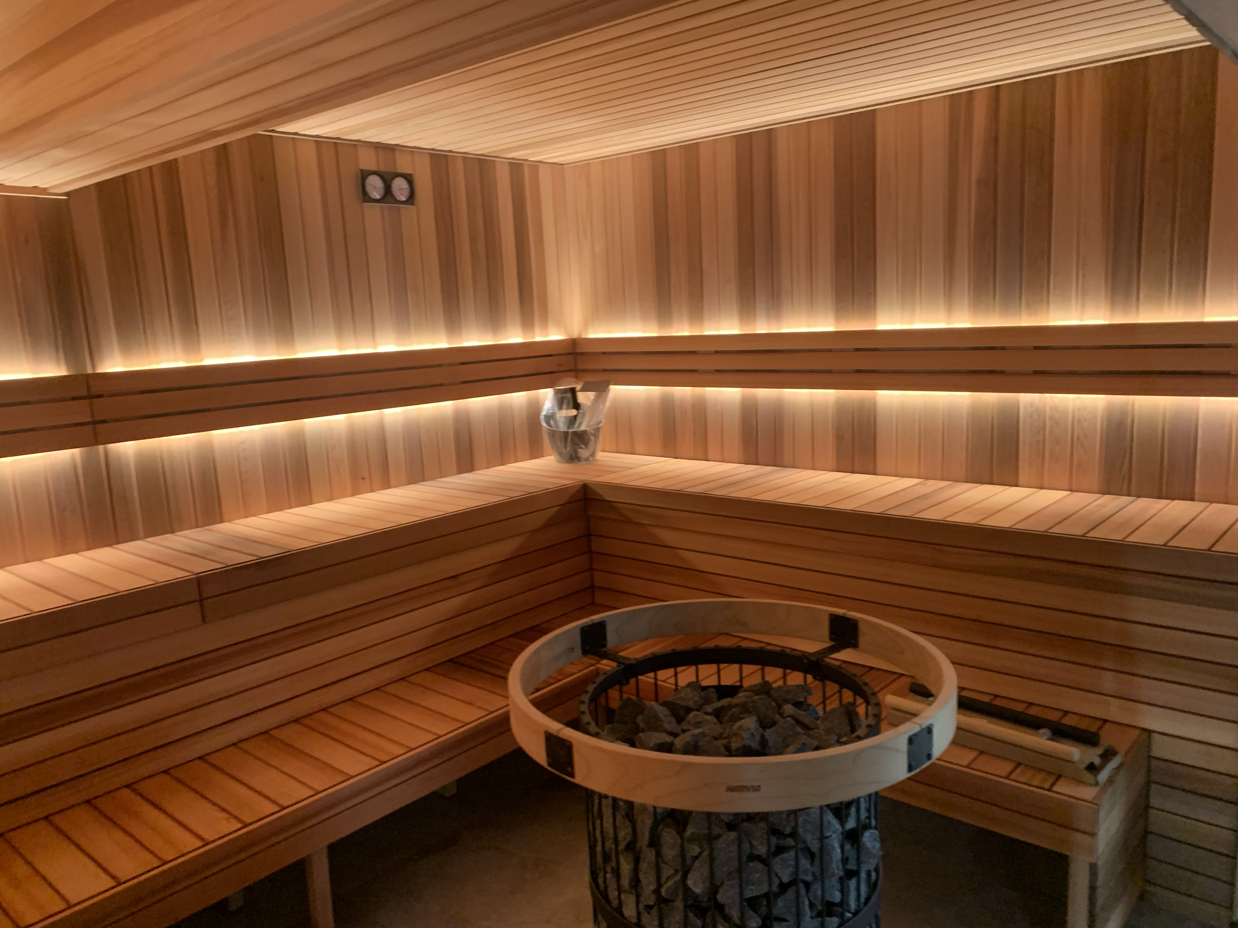 Interior of a sauna with wooden paneling, benches, and a heater with rocks, illuminated by warm lighting.