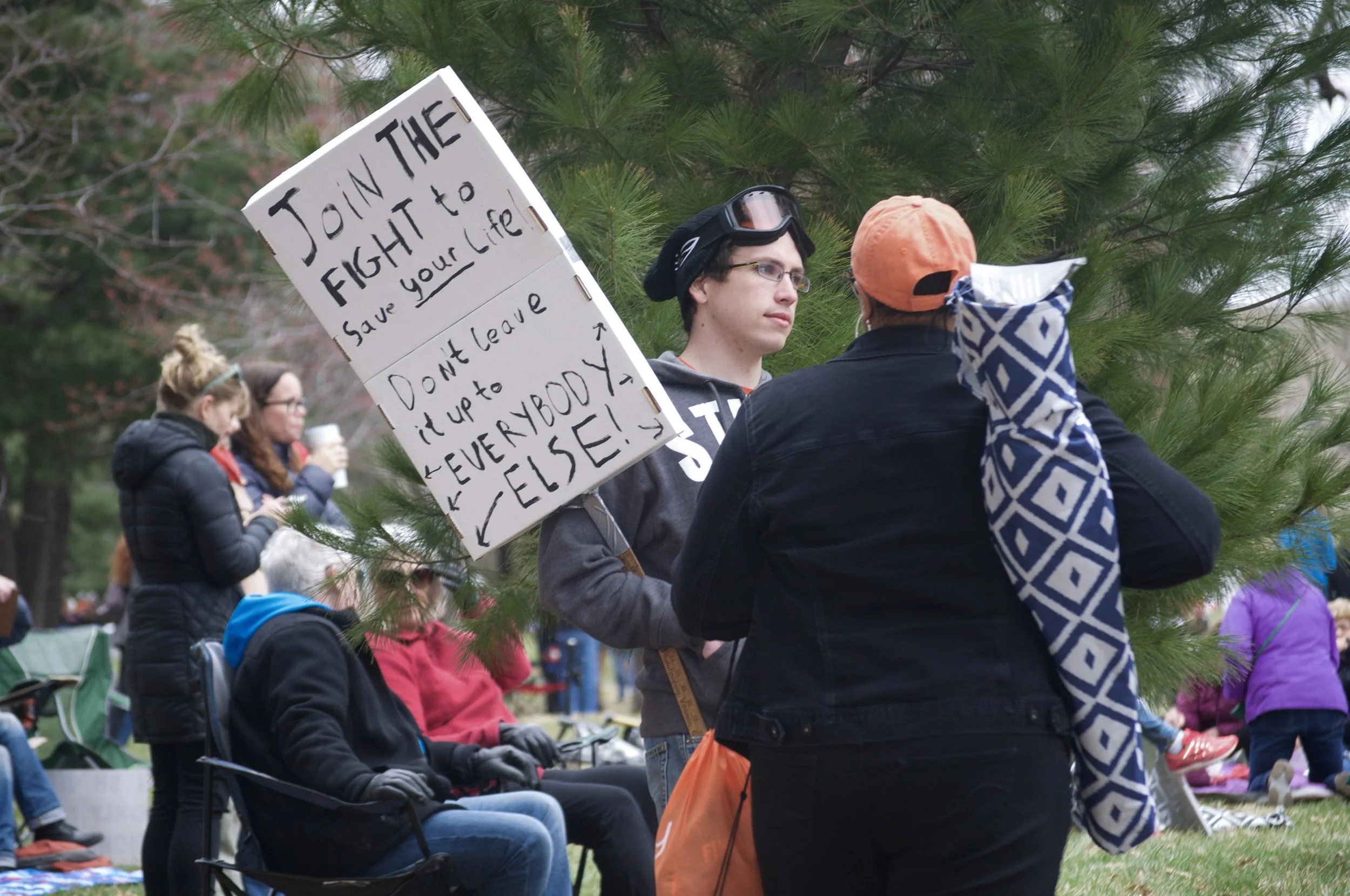 A young man holding a sign that reads 'Join the fight to save your life! Don't leave it up to everybody else!' is engaged in conversation with a woman at a public outdoor event. Several other people are visible in the background, some seated and some standing, with trees and greenery surrounding the scene.