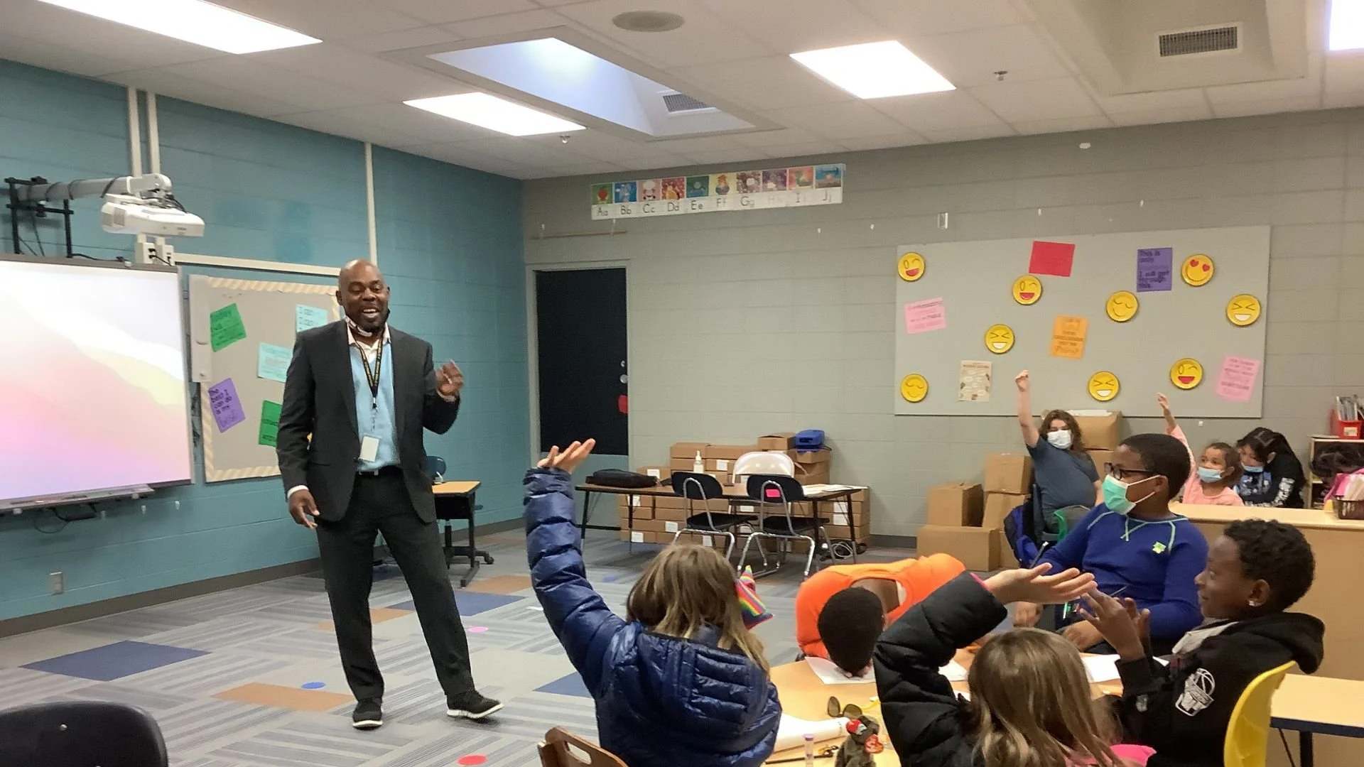 A classroom with students and a teacher. The teacher, a man in a suit, stands at the front near a whiteboard and projector. Students, some wearing masks, are seated at tables, with some raising their hands. The classroom has colorful posters and decorations on the walls.